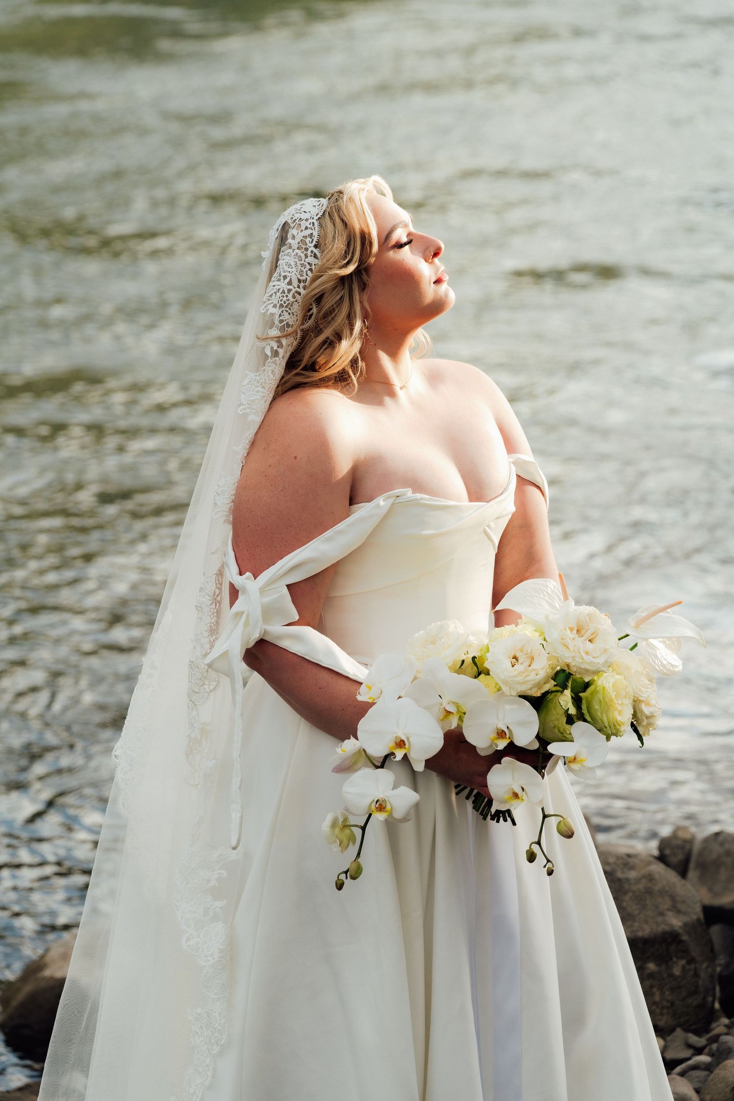 Bride in white gown holds bouquet of orchids and roses near a rocky river.