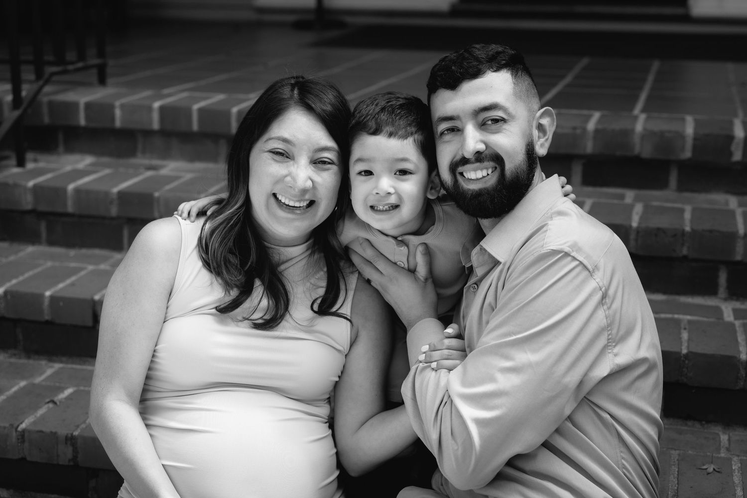 Black and white maternity photo of smiling couple with toddler sitting on brick steps.