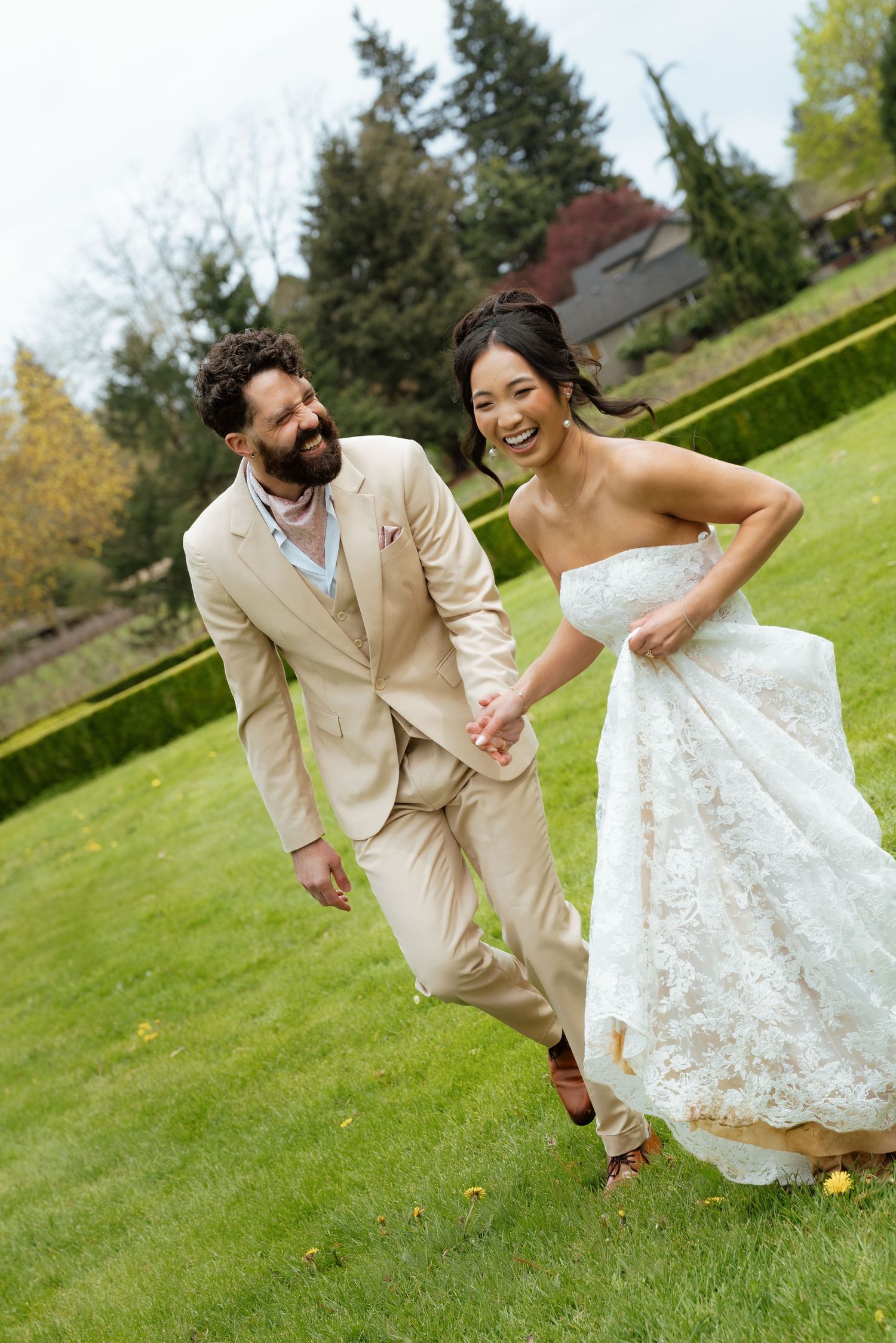 Laughing bride in strapless lace gown holds train while running with bearded groom in tan suit across green lawn.
