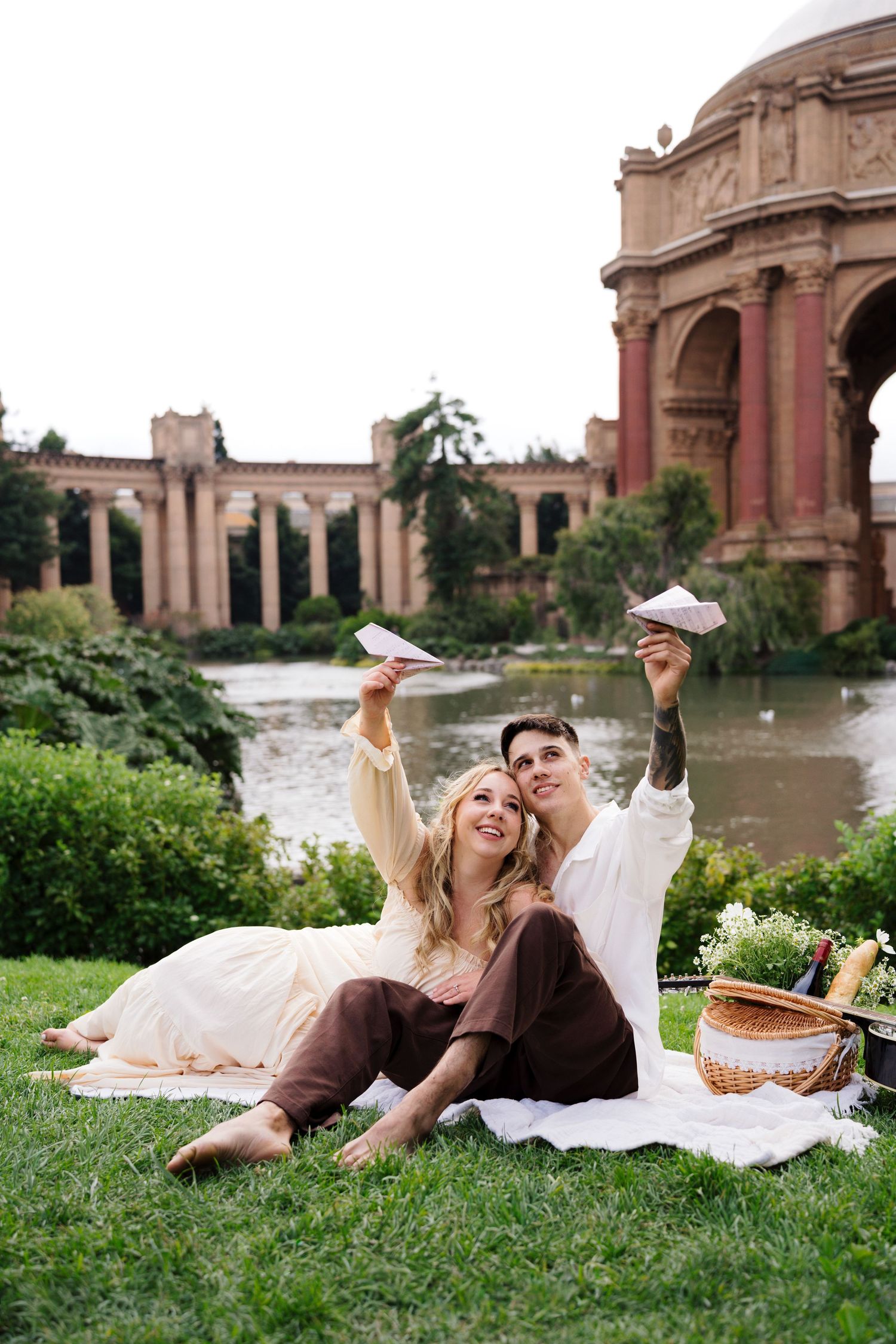 Couple sitting on blanket in park flying paper airplanes with Palace of Fine Arts in background.