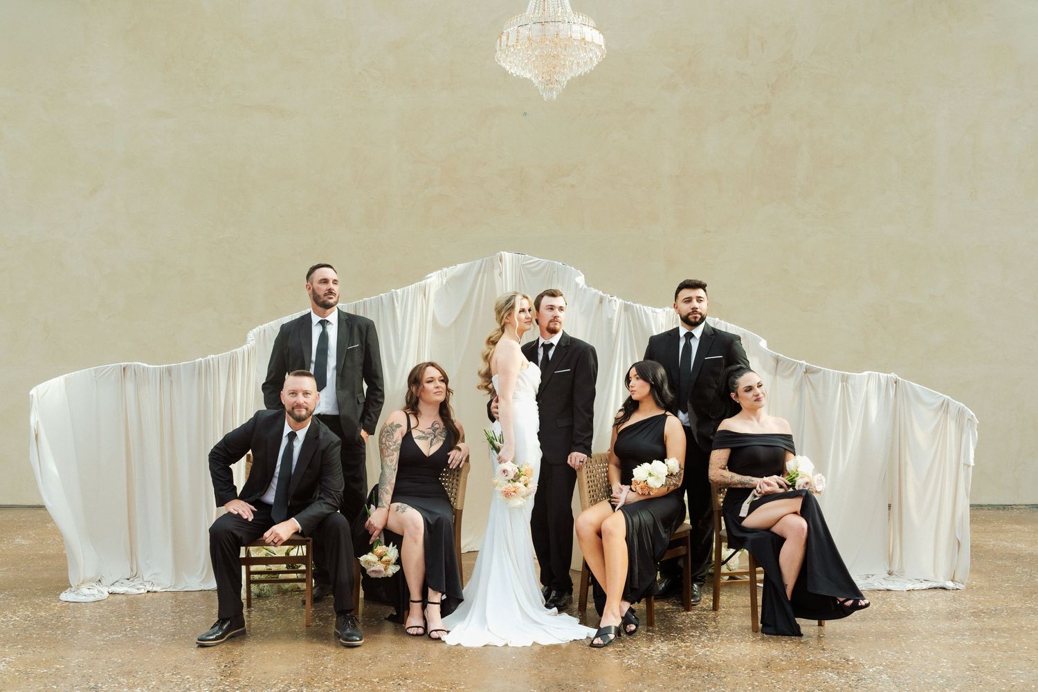 Wedding party in black attire poses elegantly with bride and groom in a decorated venue with draped fabric backdrop.