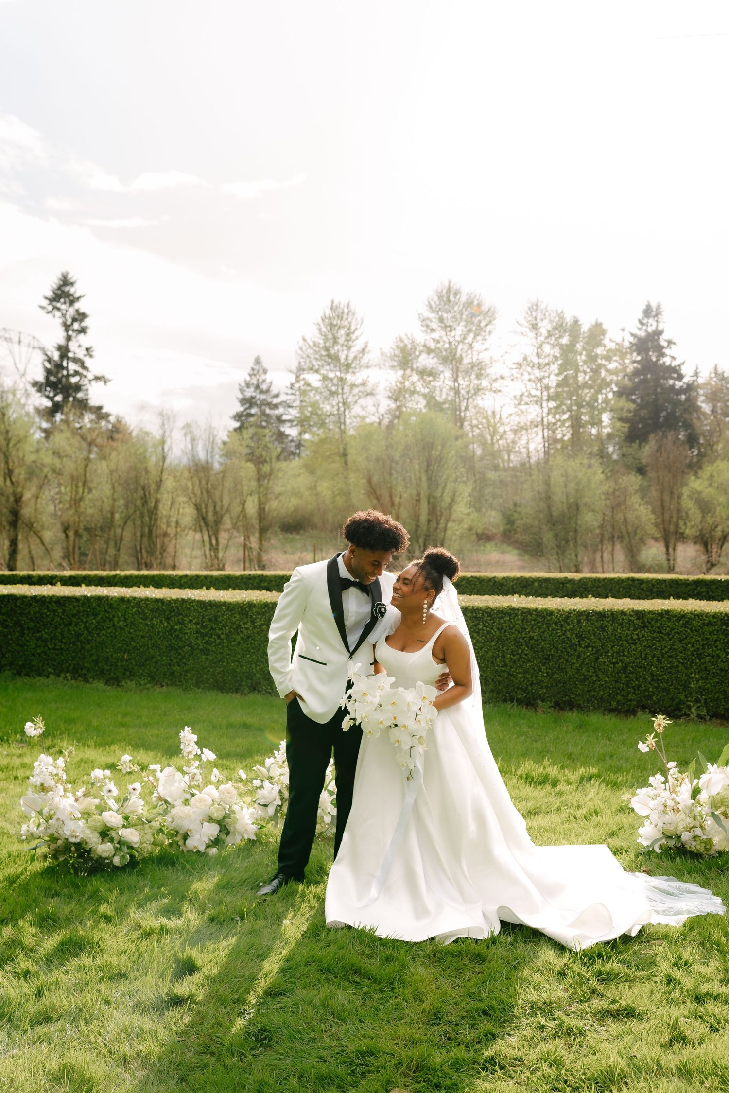 Bride and groom share a tender moment on a manicured lawn surrounded by white floral arrangements.