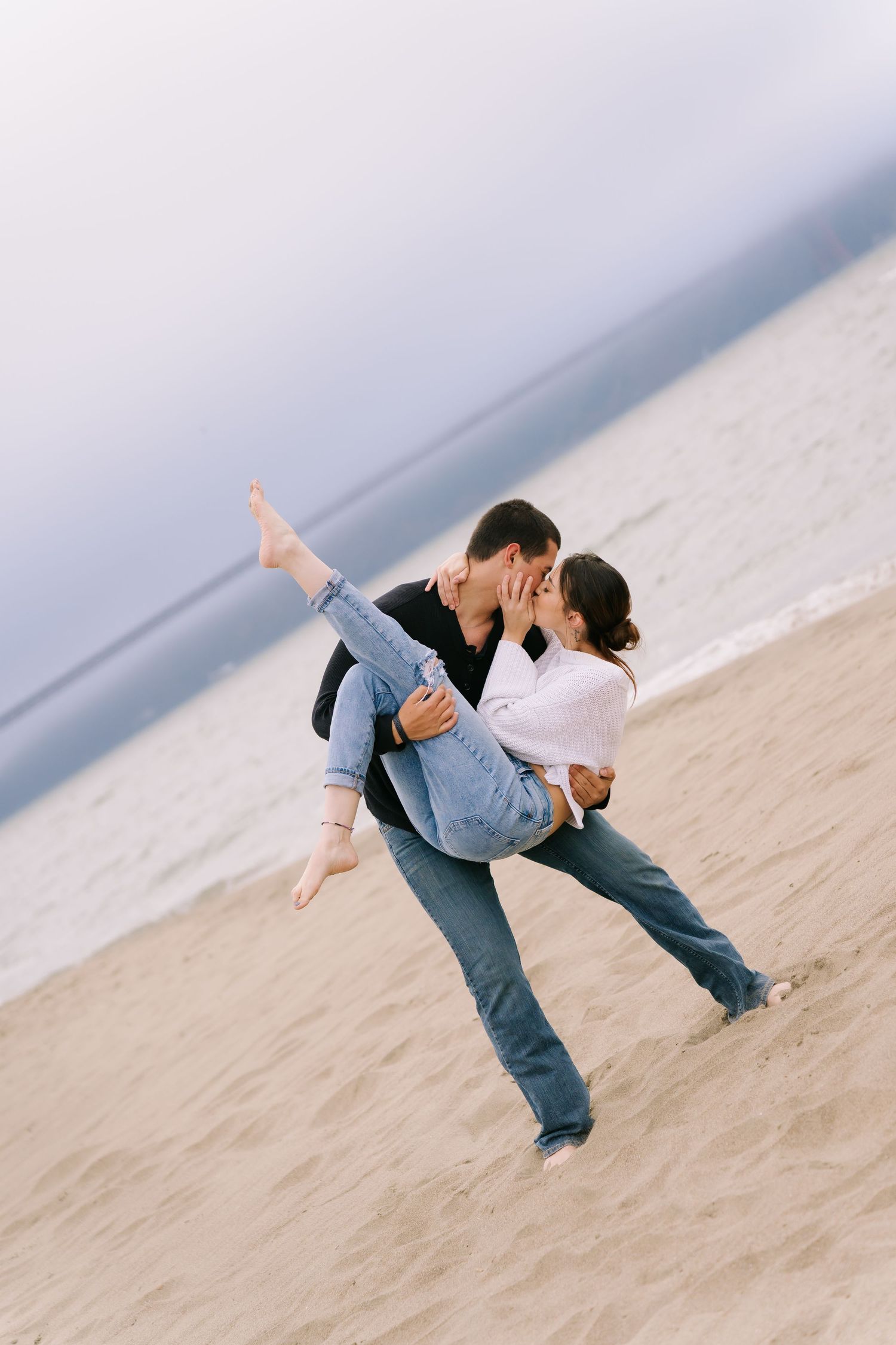 A couple shares a romantic kiss on a sandy beach, as the man lifts the woman in his arms near the ocean.