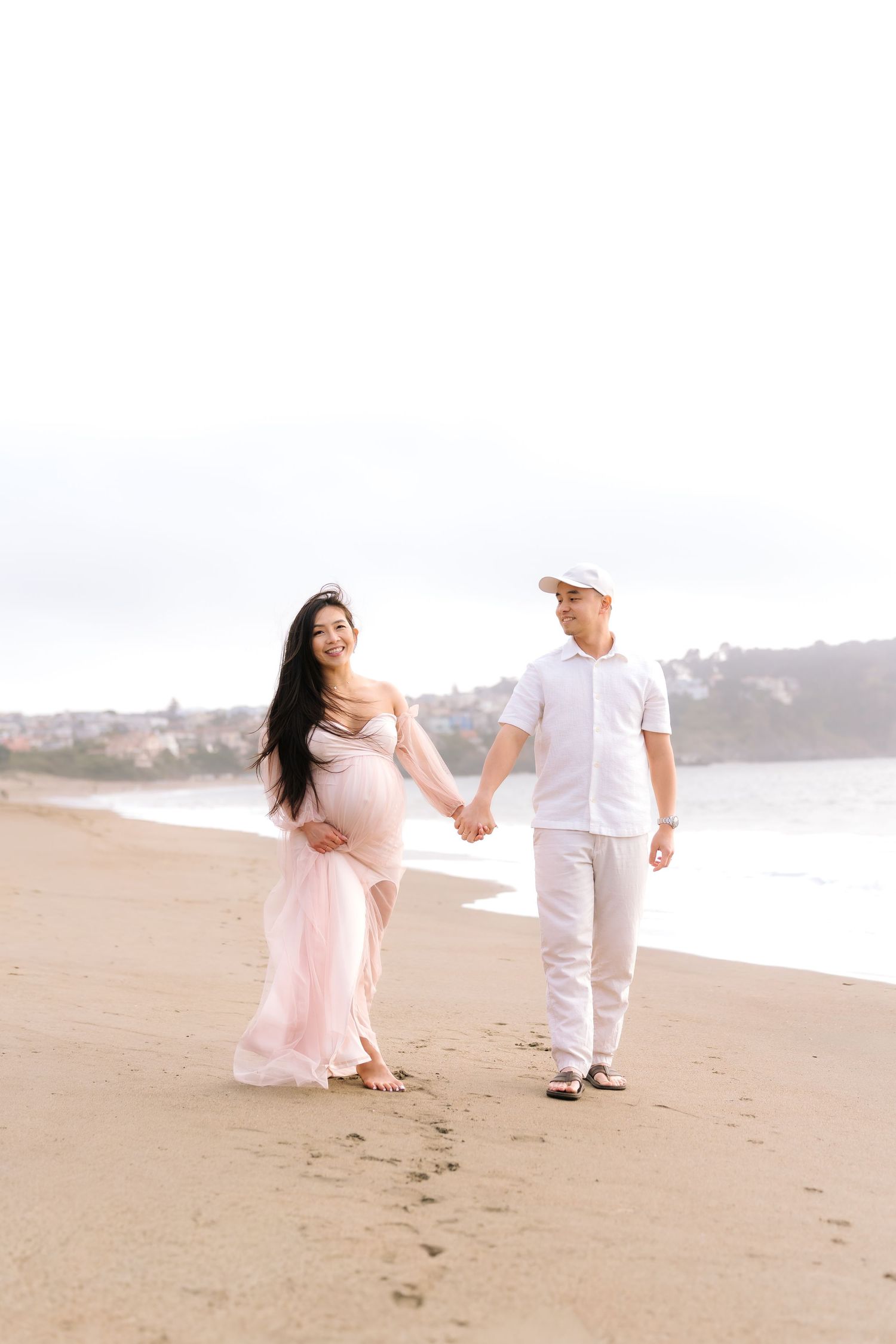 Pregnant couple walking hand-in-hand on a sandy beach, woman in pink maternity gown, man in white outfit.