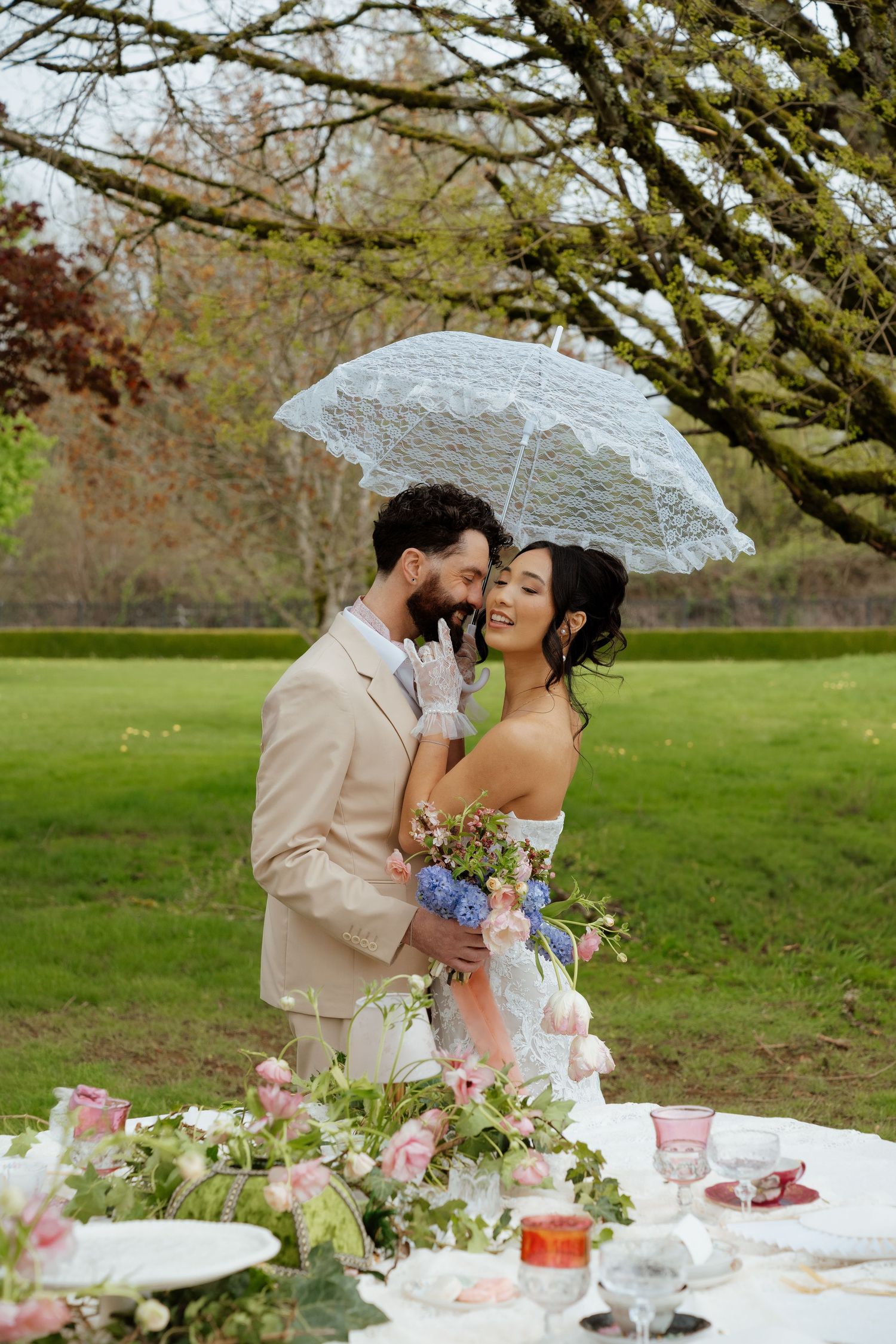 Bride and groom share tender moment under white lace umbrella at outdoor garden wedding table.