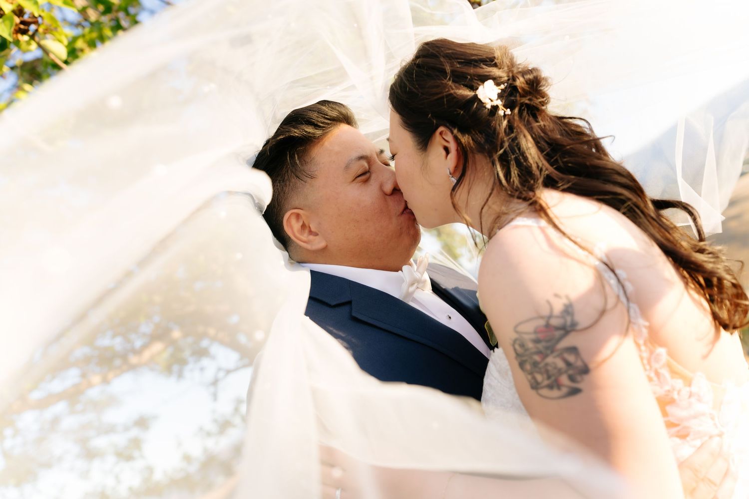 Bride and groom share a romantic kiss under a flowing white veil outdoors on their wedding day.