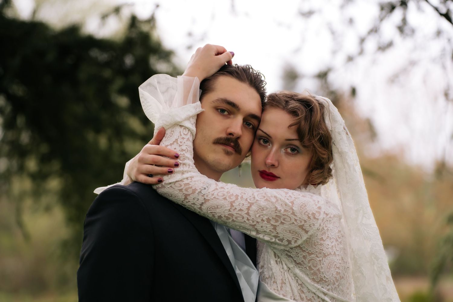 Vintage-styled bride in lace veil embraces groom with mustache in black suit outdoors.