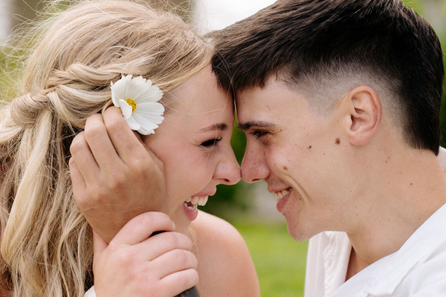 A young couple smiling and touching foreheads, the woman wearing a white flower in her hair.