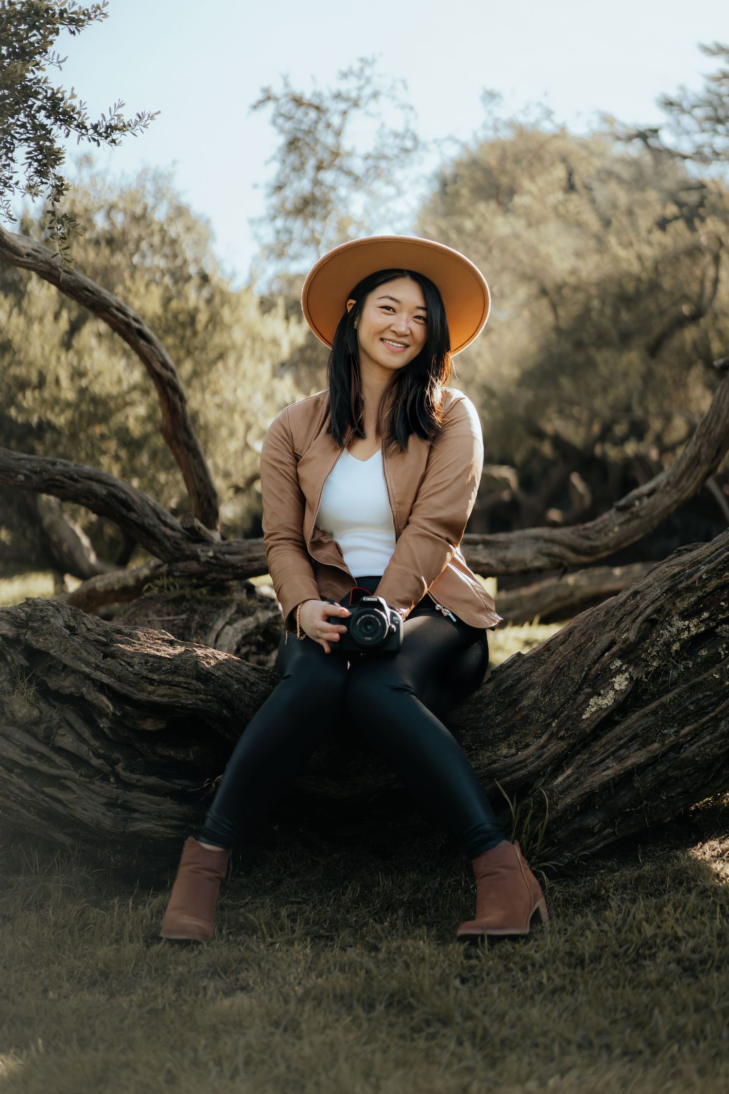 Smiling woman in orange hat holding camera sitting on twisted tree trunk in outdoor park setting.