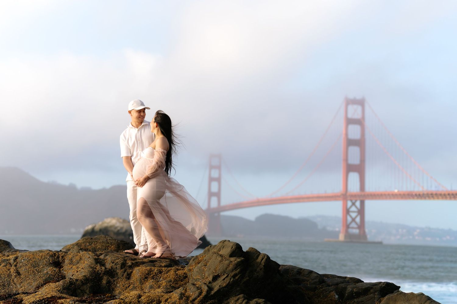 Couple embracing on rocks with Golden Gate Bridge backdrop in San Francisco, wearing white outfits against a misty sky.