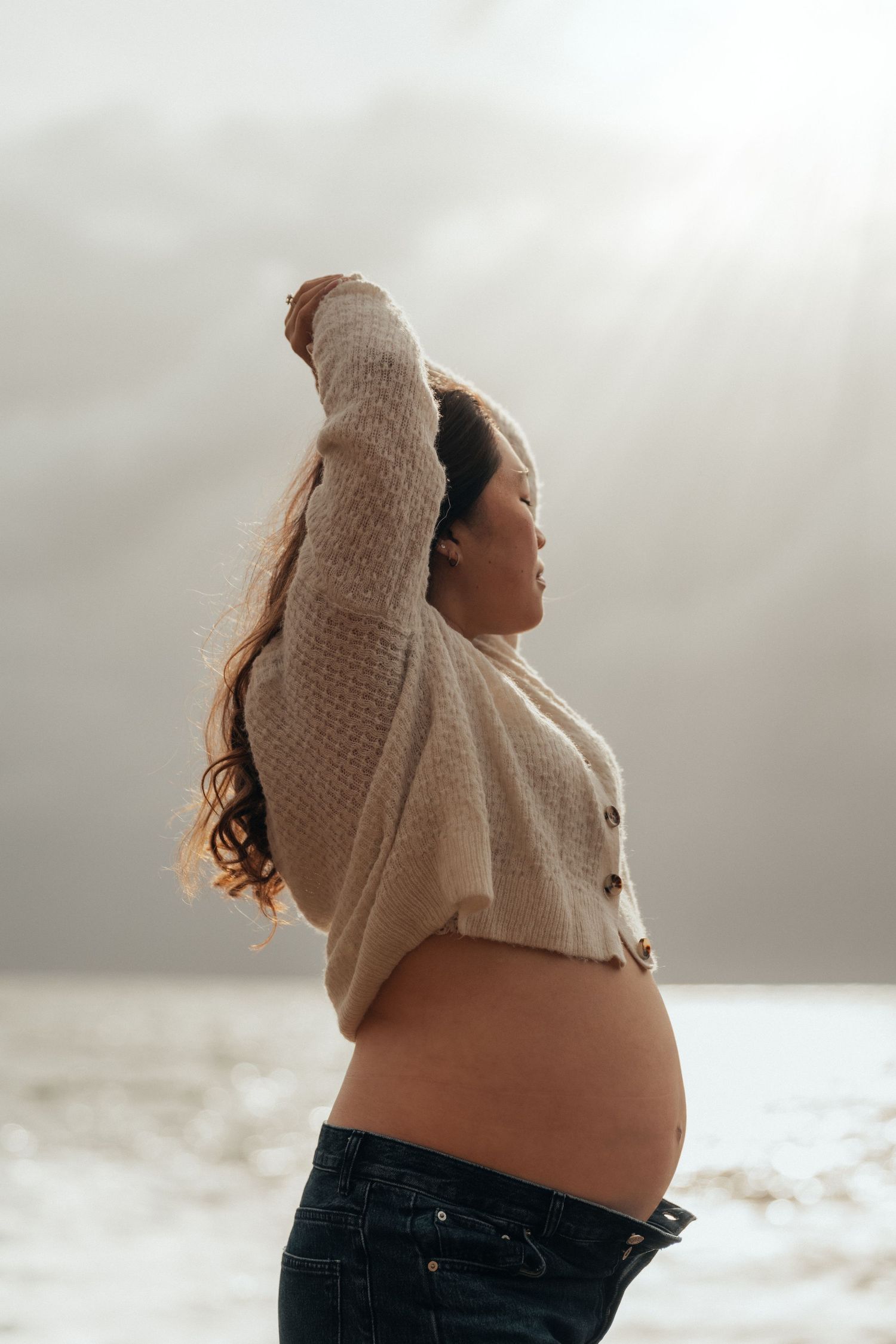 Pregnant woman in cropped beige sweater and jeans poses outdoors in snowy landscape with arm raised.