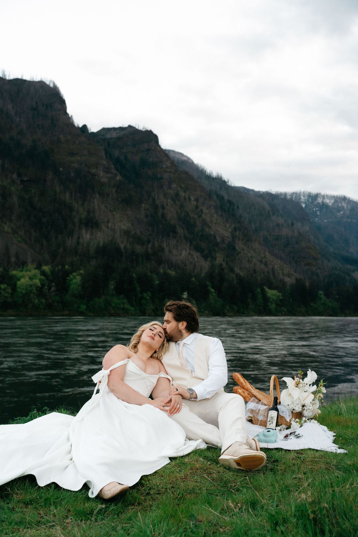 Couple relaxes on picnic blanket by river with mountain backdrop on wedding day.