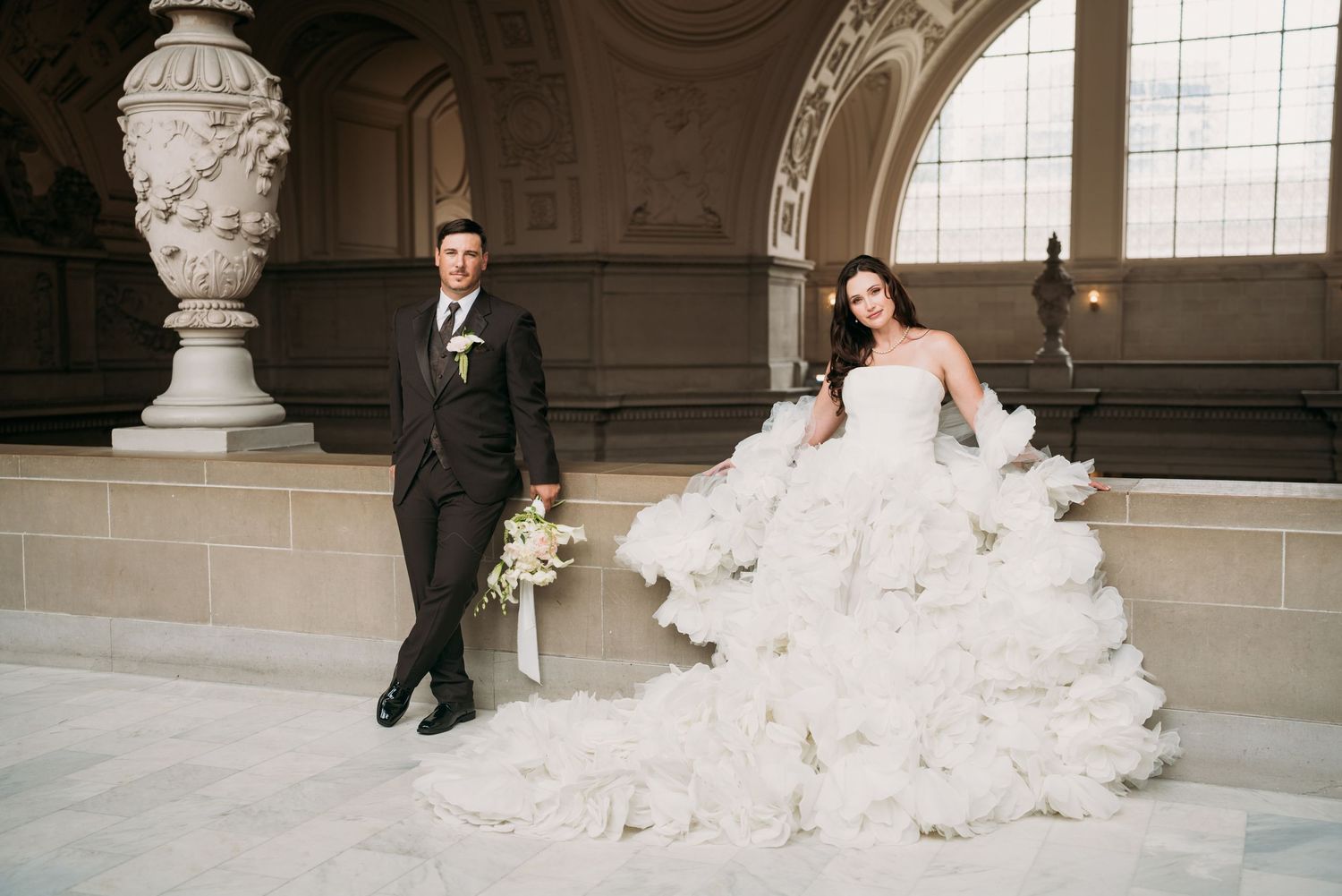 Bride in voluminous white ruffled gown and groom in black tuxedo posing elegantly in a grand marble wedding venue.