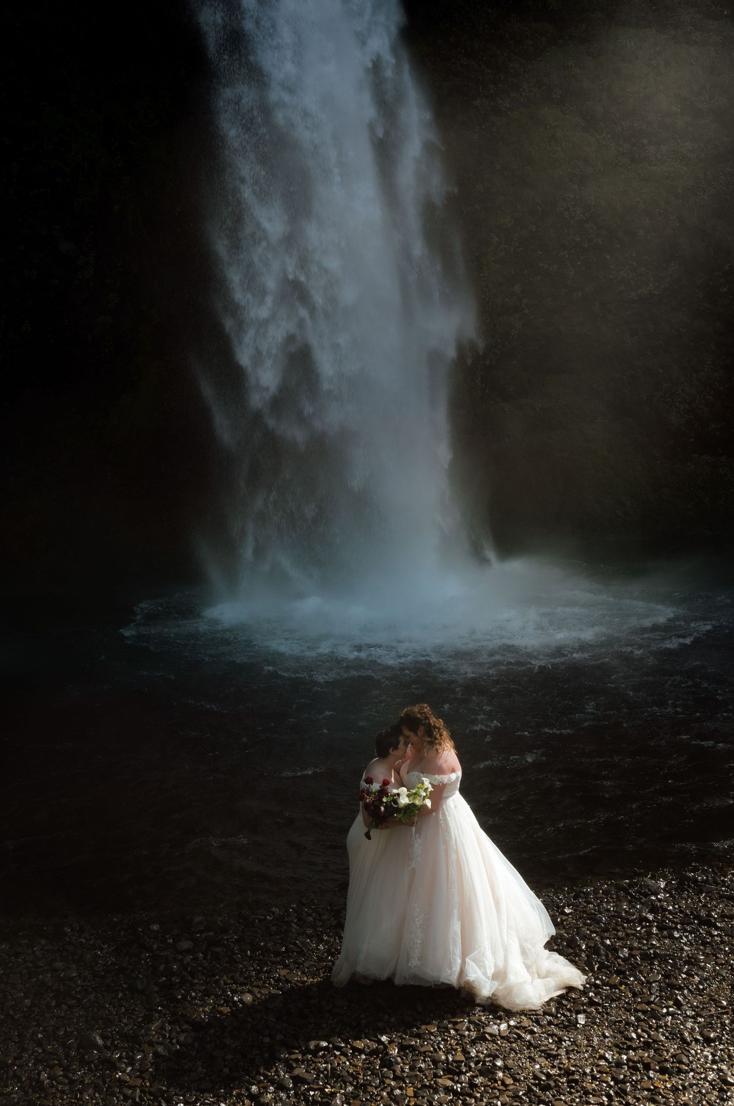 Bride in white gown holds bouquet before misty waterfall in dramatic dark setting.