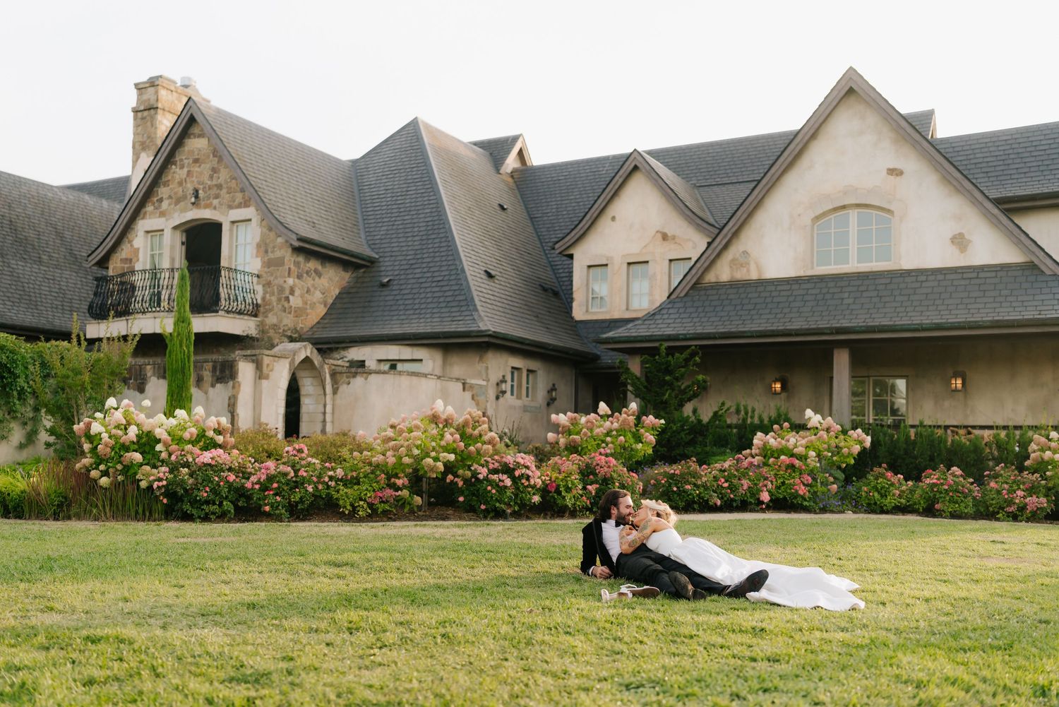Bride and groom pose romantically on green lawn before elegant stone manor with blooming pink roses.