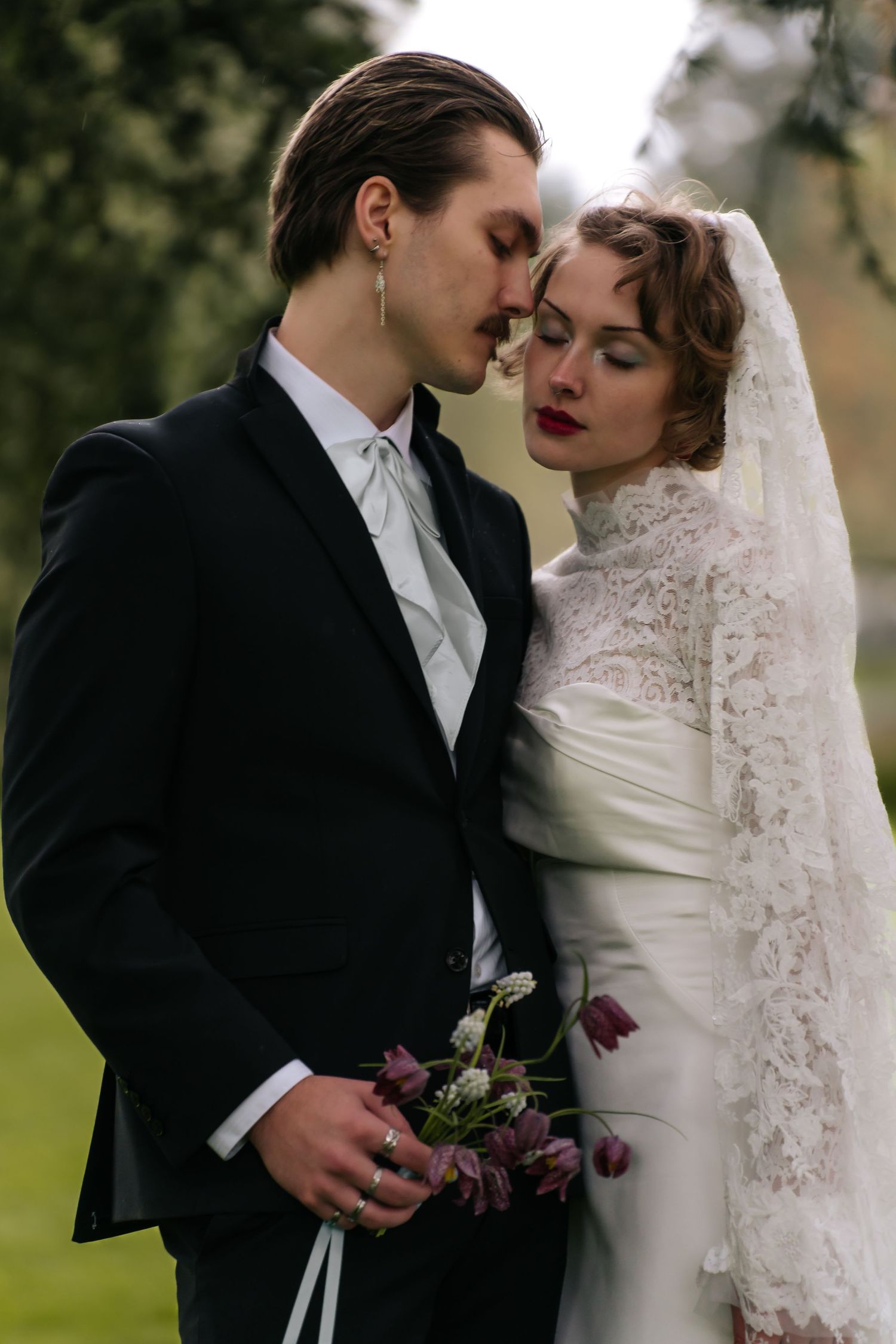 Elegant wedding couple in formal attire sharing an intimate moment outdoors, bride in lace veil holding purple bouquet.