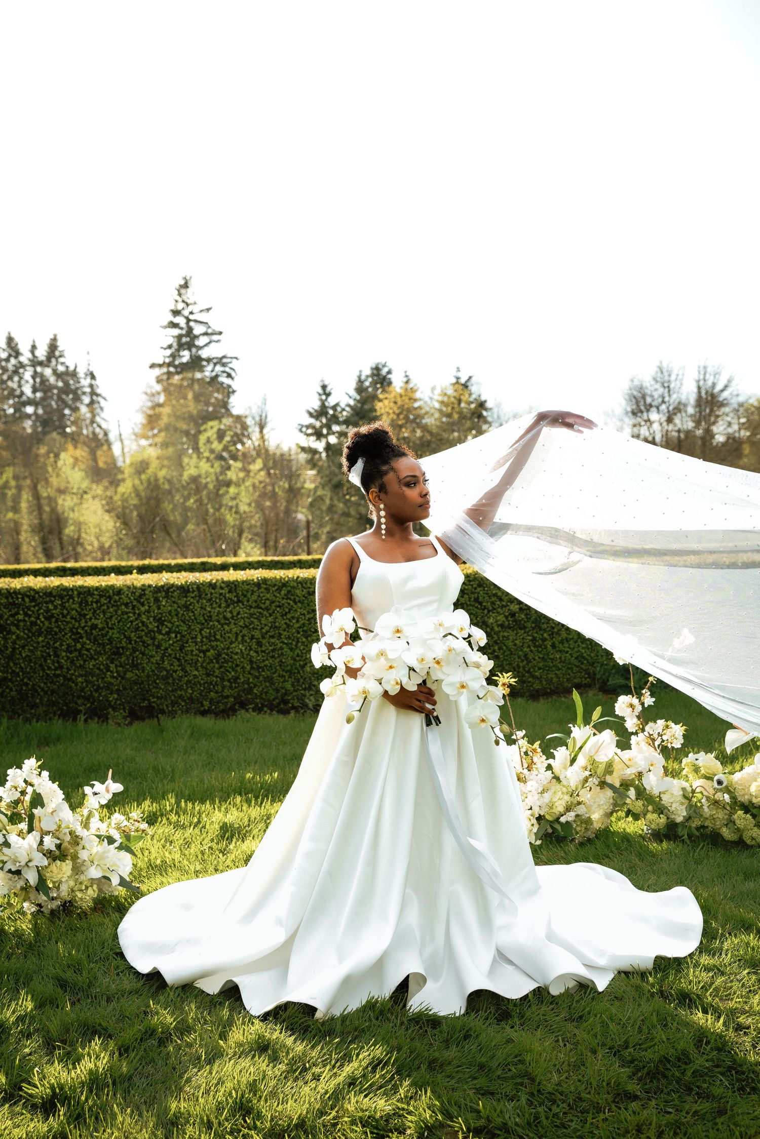 Black bride in white ballgown tosses veil in sunlit garden with white florals.
