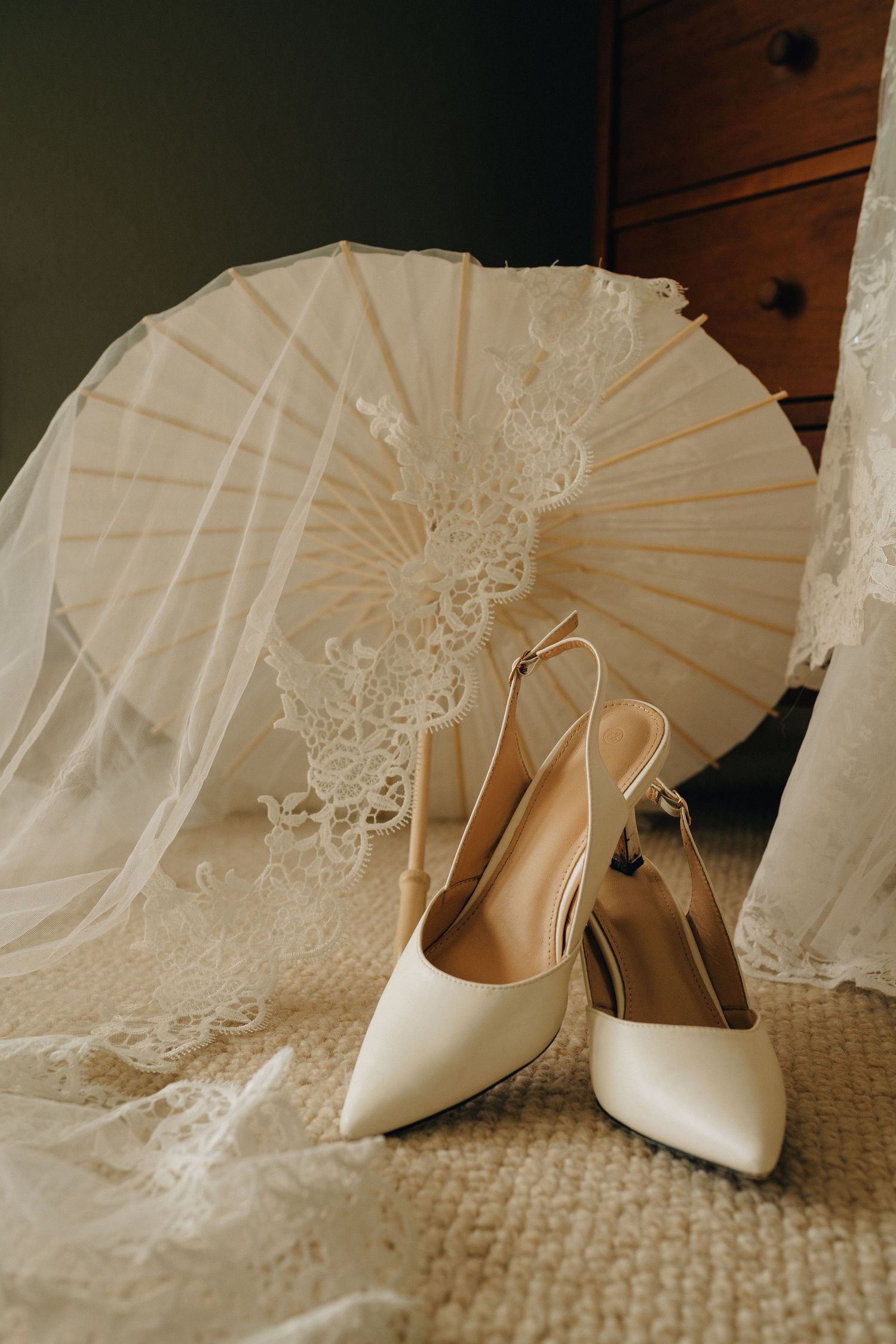 White satin slingback bridal heels displayed beside lace parasol and flowing veil on textured carpet.