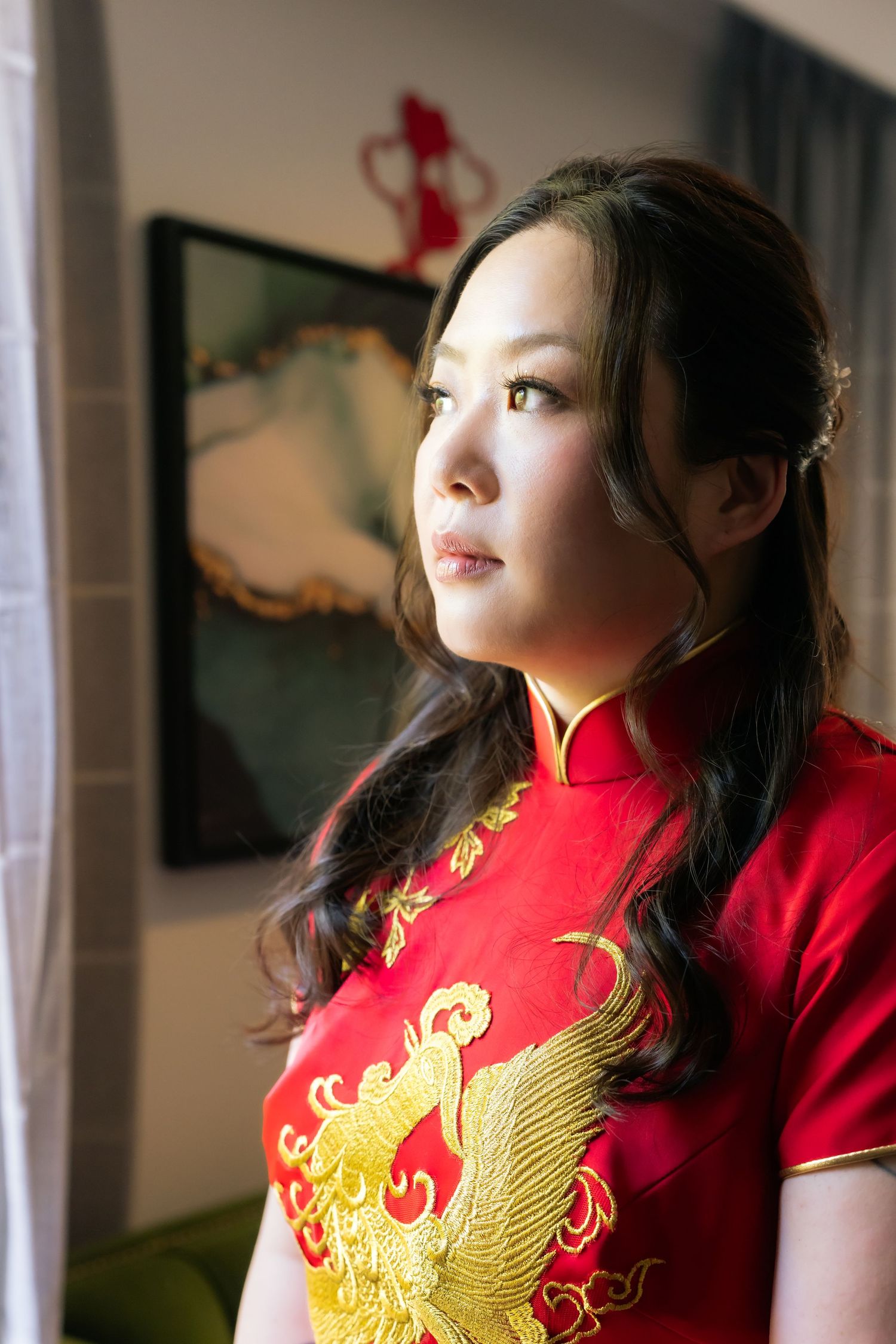 Young Asian woman in red and gold embroidered cheongsam qipao dress gazing thoughtfully near a window.