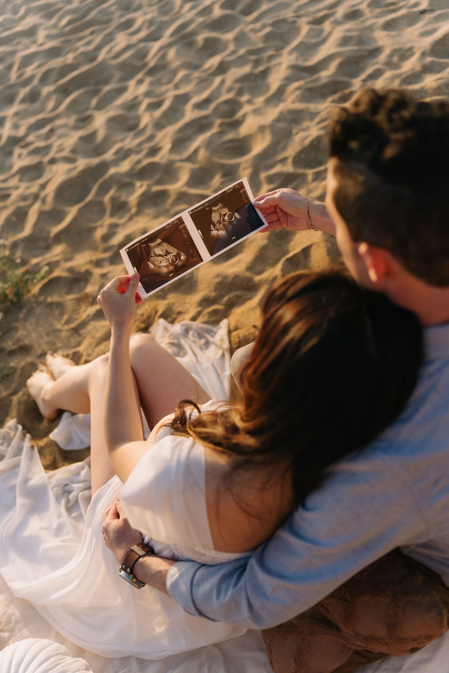 Expectant couple sitting on a sandy beach at sunset, holding ultrasound photos of their baby while embracing.