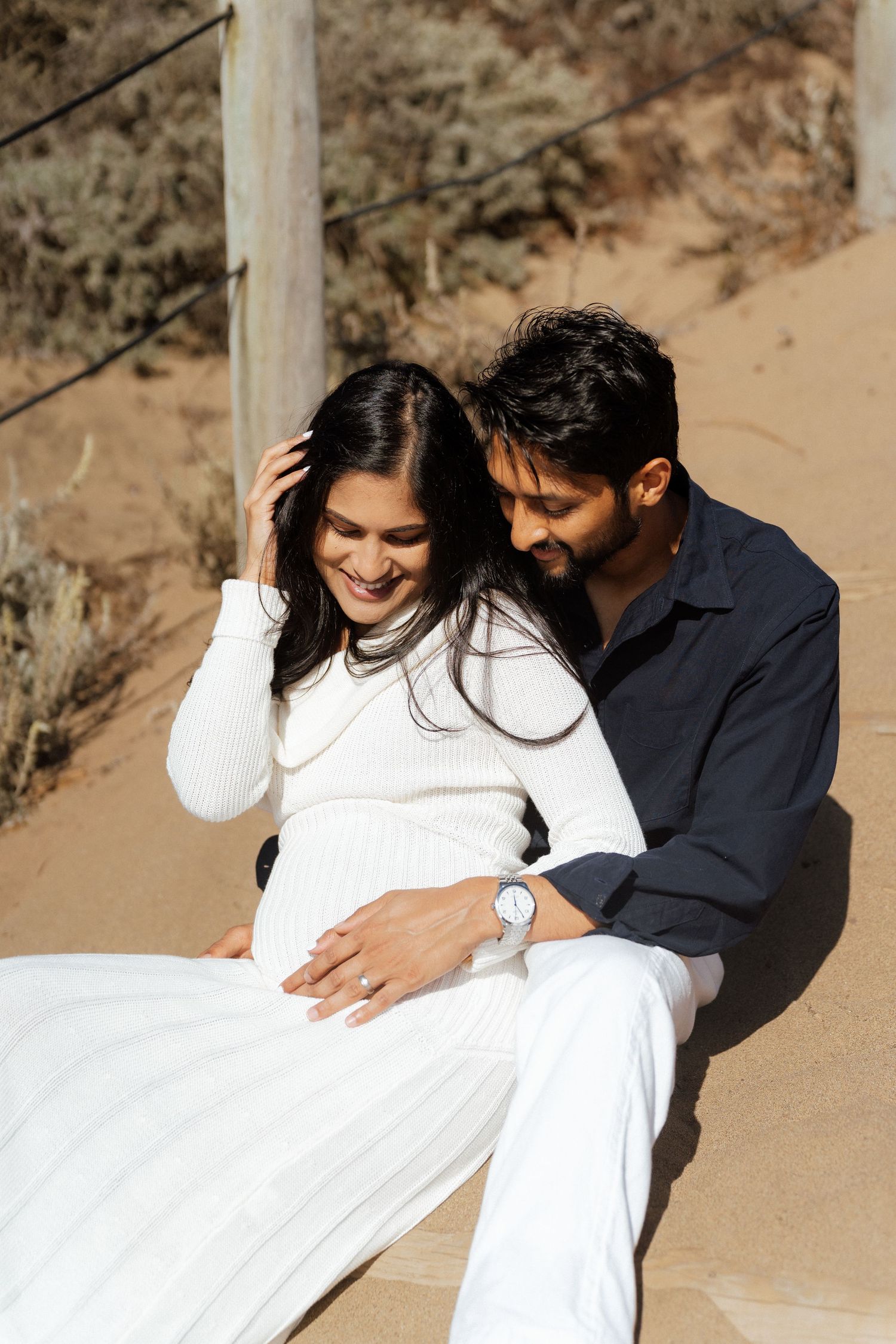 A couple sits on sandy dunes, woman in white dress smiling as man in navy shirt embraces her tenderly.