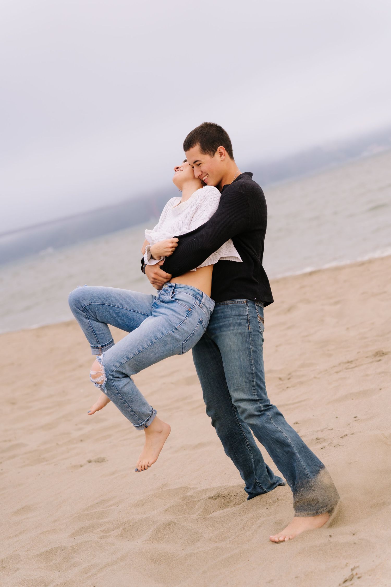 Romantic couple embracing on a sandy beach, man lifting woman in jeans and white top near the ocean.