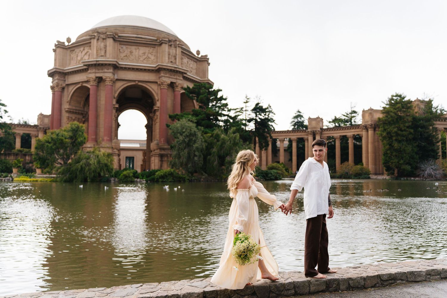 Bride and groom holding hands at the Palace of Fine Arts in San Francisco with a lagoon backdrop.