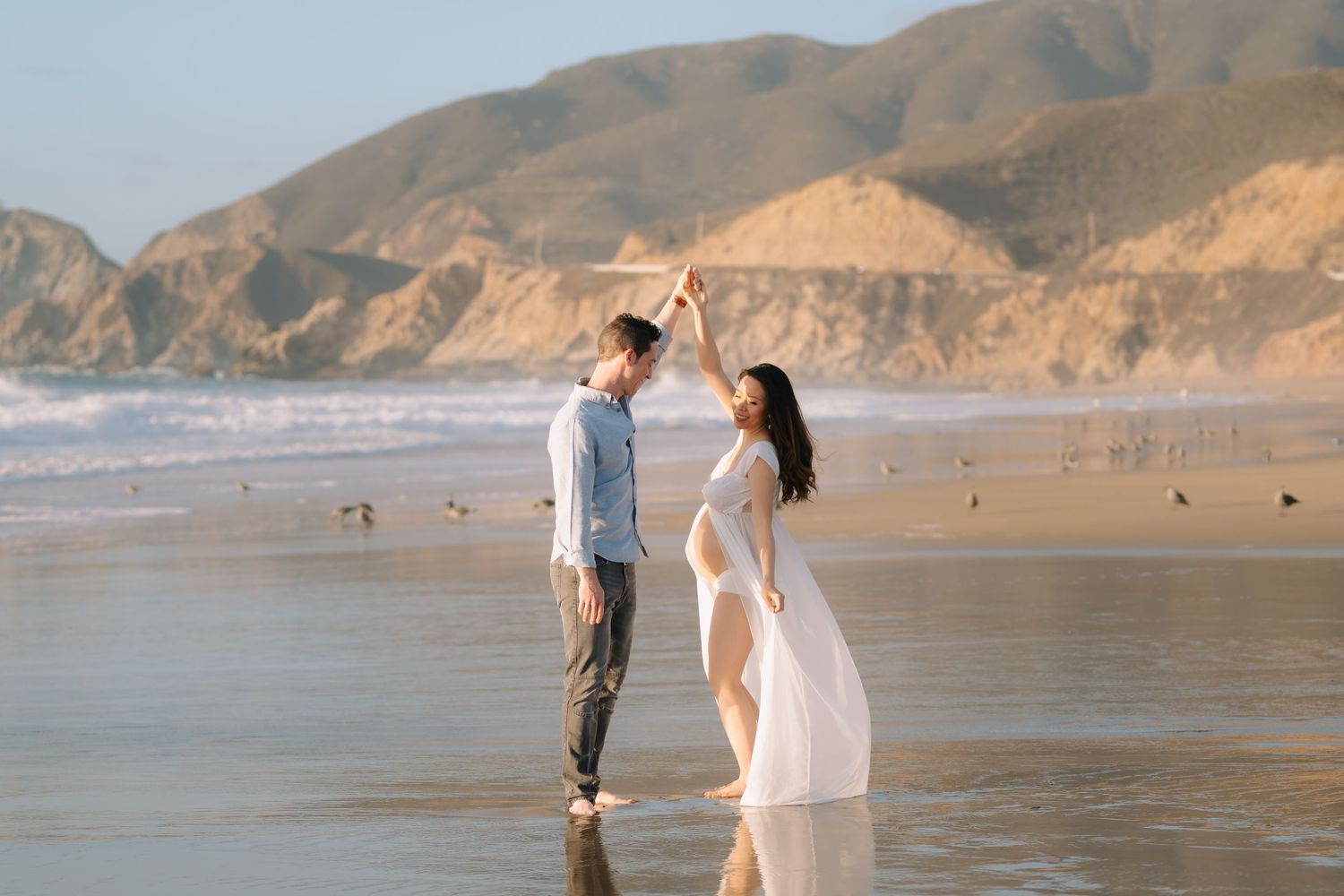 Pregnant woman in a flowing white gown twirls under her partner's raised hand during a maternity photoshoot on a wet sandy beach with hills.