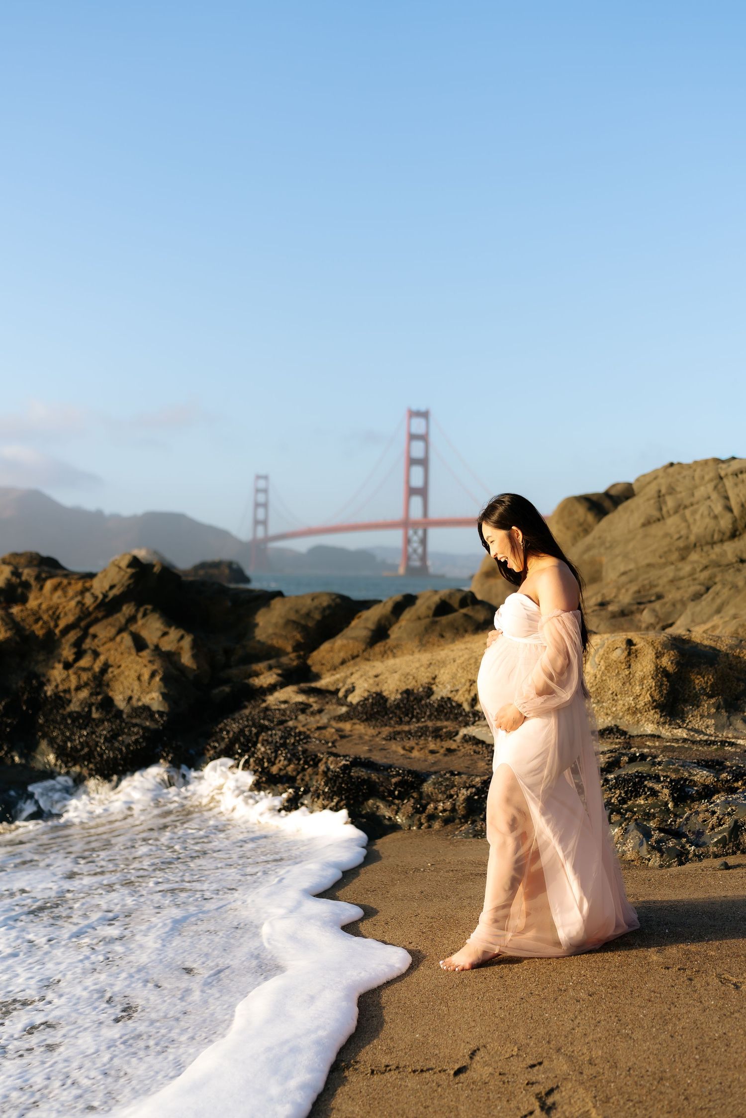 Pregnant woman in pink gown stands on Baker Beach with Golden Gate Bridge backdrop in San Francisco.