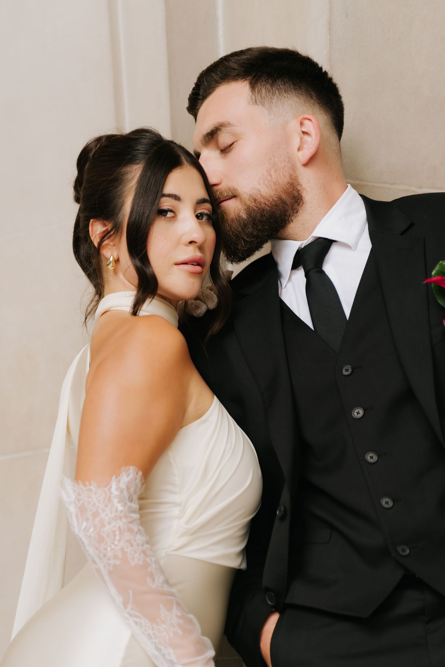 A bride in a white gown and veil leans against a groom in a black suit as he gently kisses her temple.