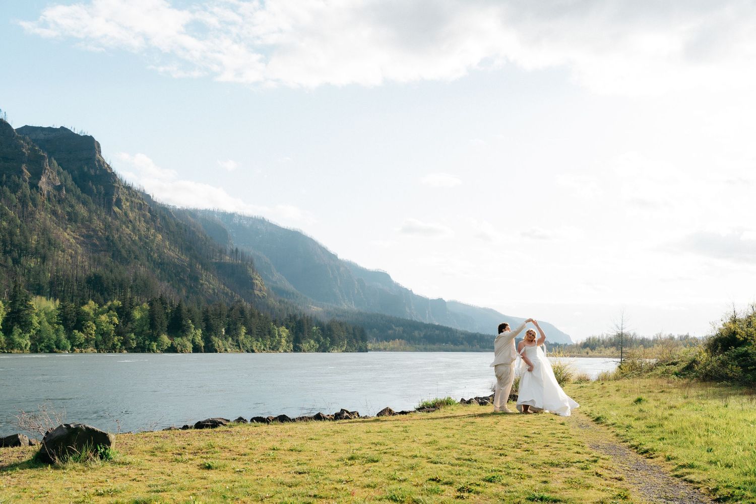 Couple embracing in wedding attire by a scenic river with mountain backdrop and lush greenery under soft sunlight.