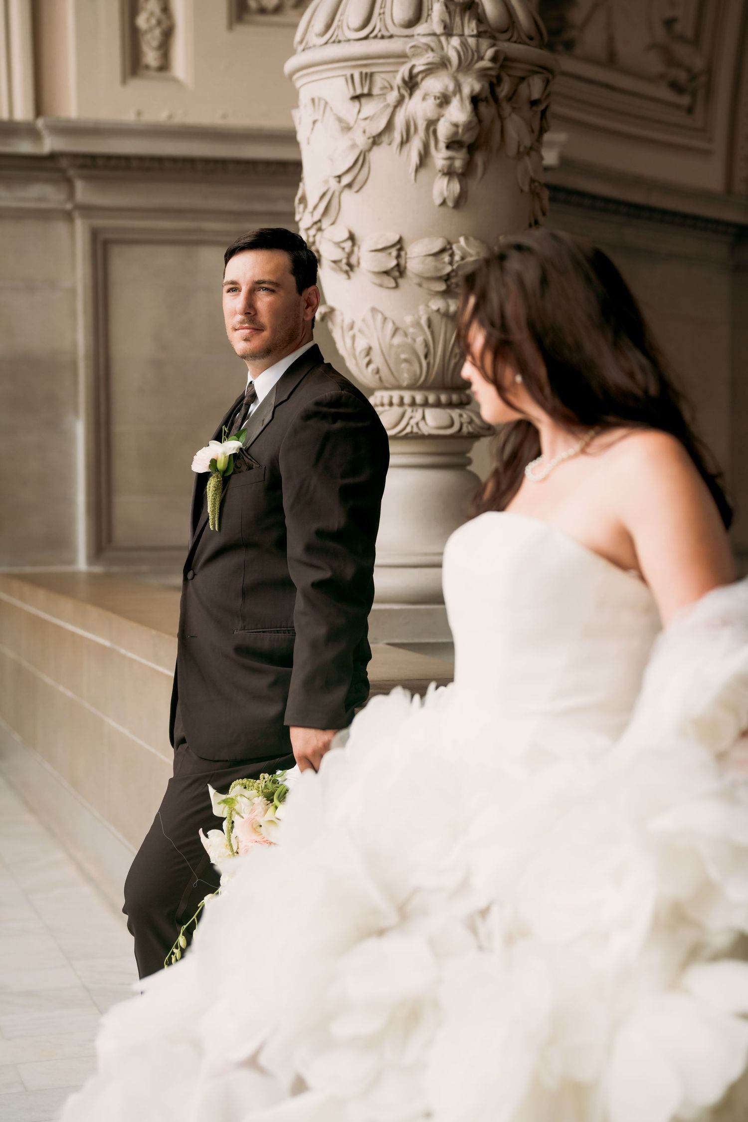Groom in dark suit stands by ornate column as bride in ruffled white gown poses in foreground at elegant venue.