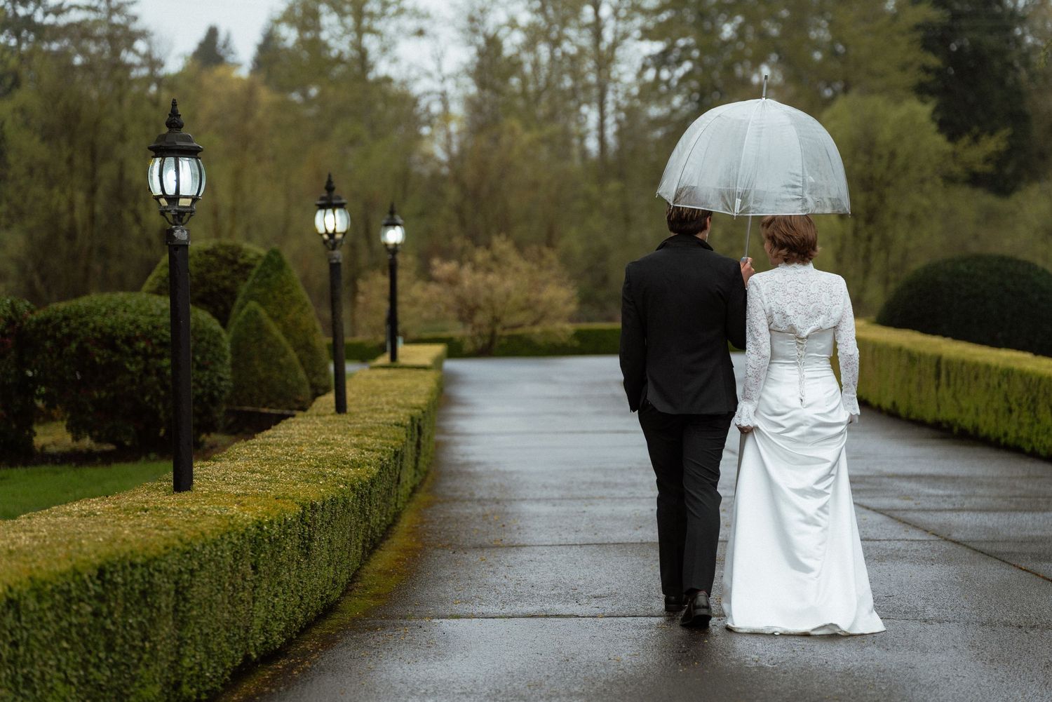 Newlywed couple walks down a rainy garden path sharing a clear umbrella.