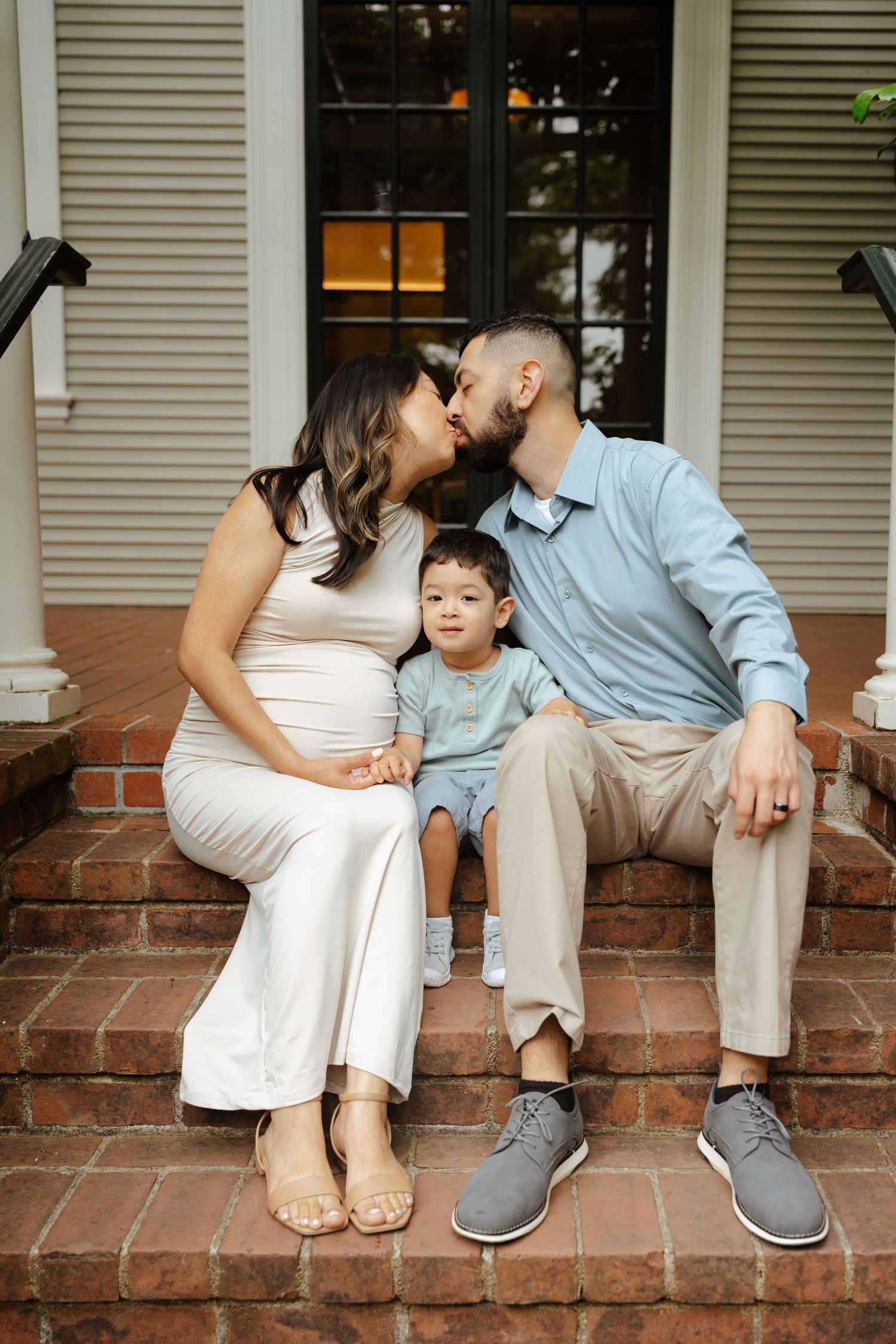 Pregnant woman and man kissing on brick steps with toddler between them during outdoor family maternity photo session.