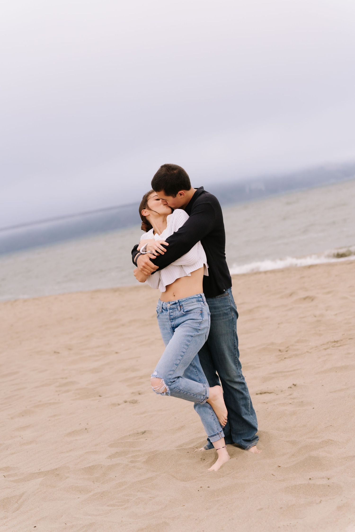 Couple embracing and kissing on a sandy beach with calm ocean waves in the background on an overcast day.