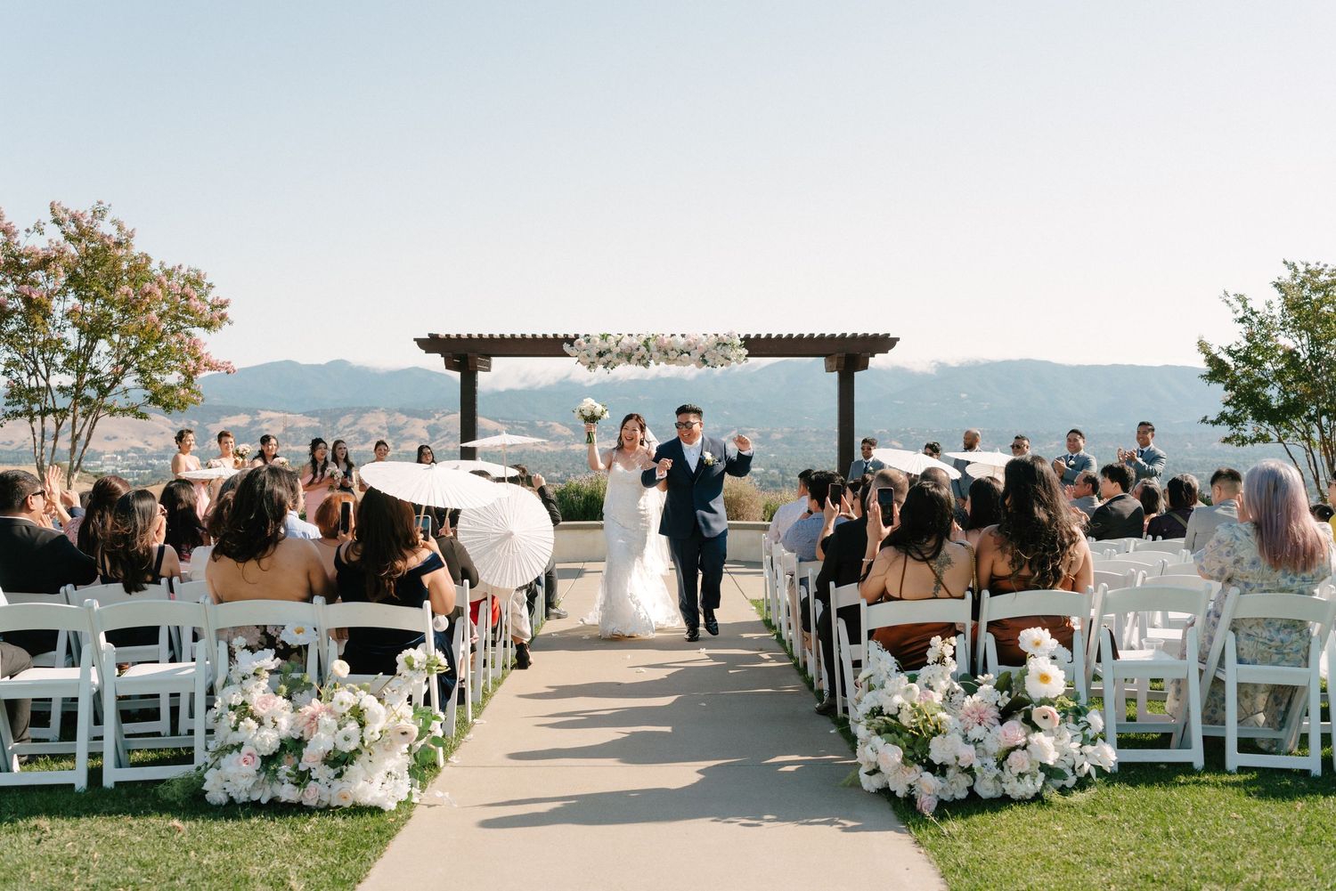 Newlyweds walk down outdoor aisle flanked by white floral arrangements and guests at a sunny rooftop wedding ceremony.