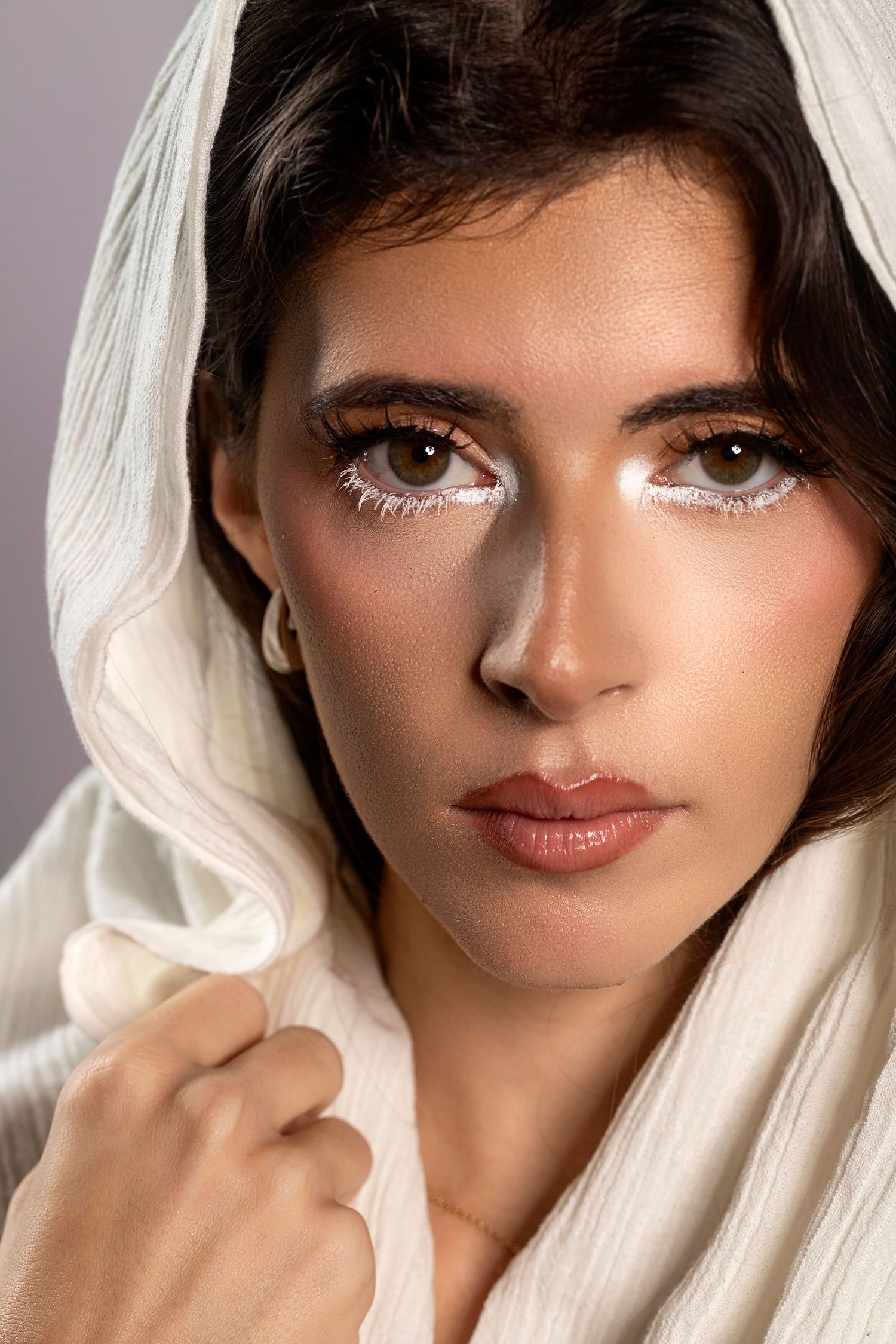Close-up portrait of a young woman wearing a white headscarf with artistic white eyeliner makeup against a gray background.