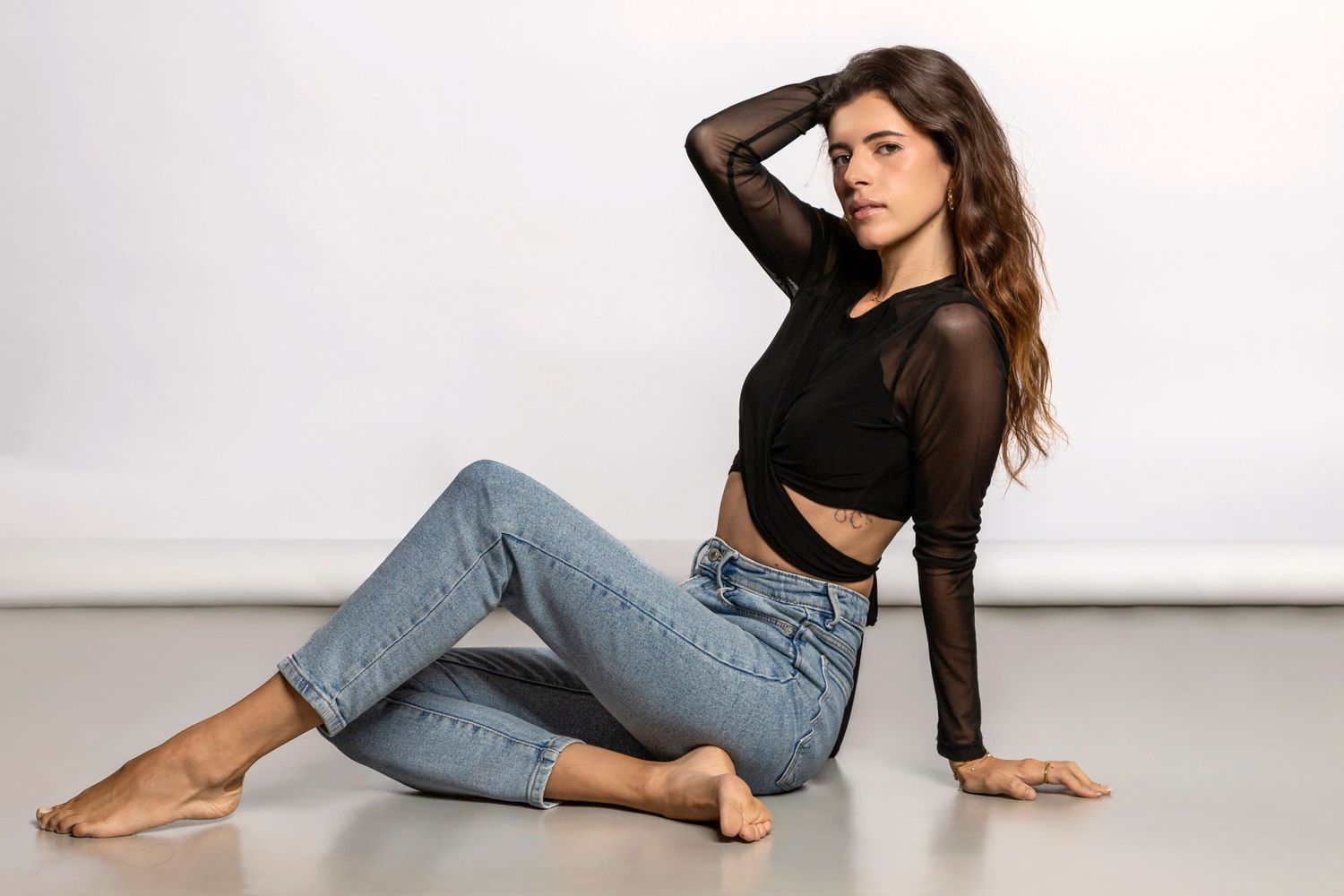 Young woman in black crop top and jeans sitting on floor, posing barefoot against white background.