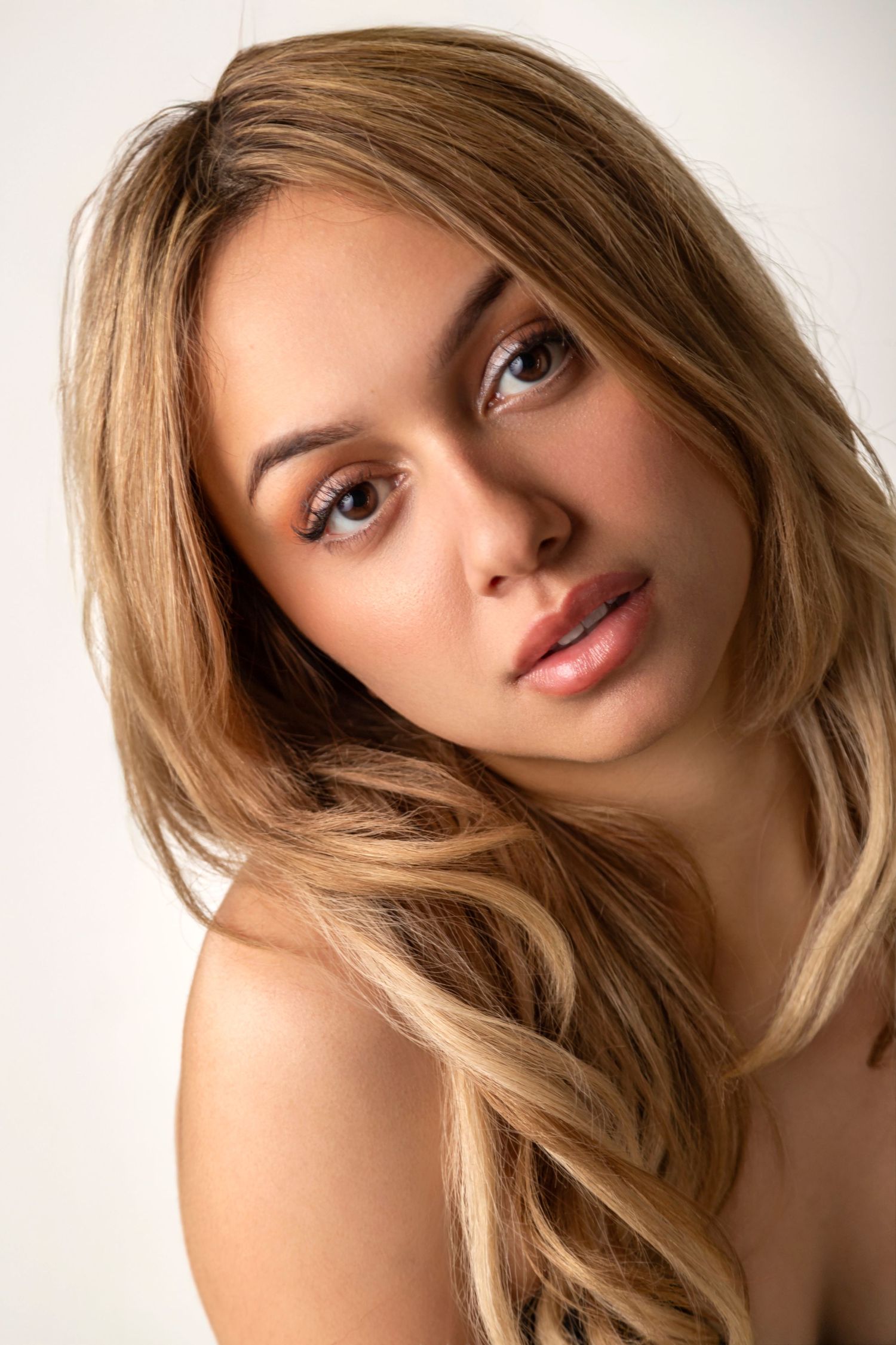 Young woman with long blonde wavy hair and natural makeup poses against a light background.