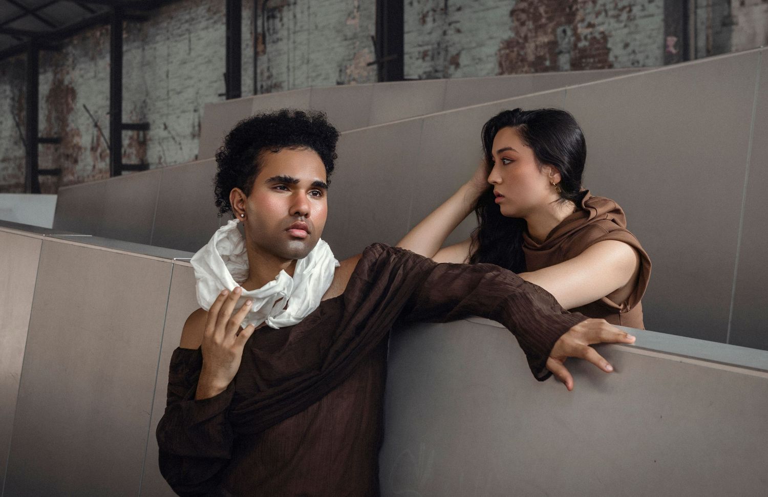Two young models pose dramatically in brown draped garments against industrial staircase in a fashion editorial.