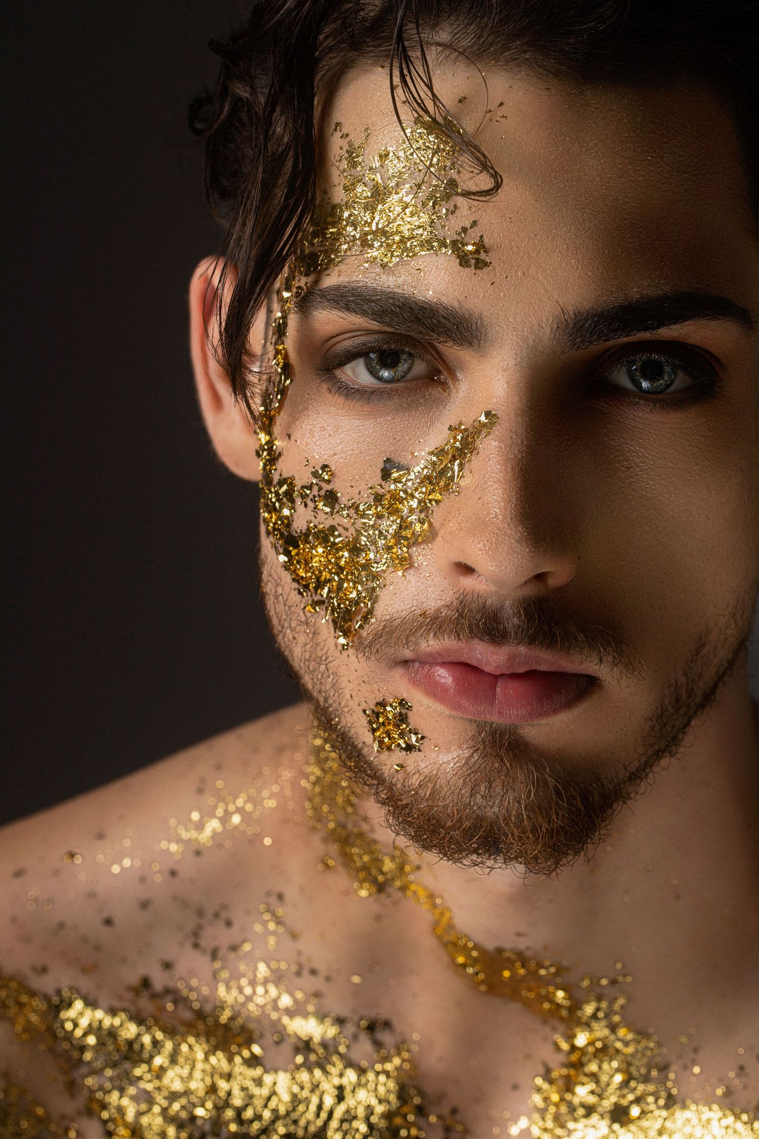 Young man with blue eyes and gold glitter decoratively applied to his face, beard, and chest against a dark background.