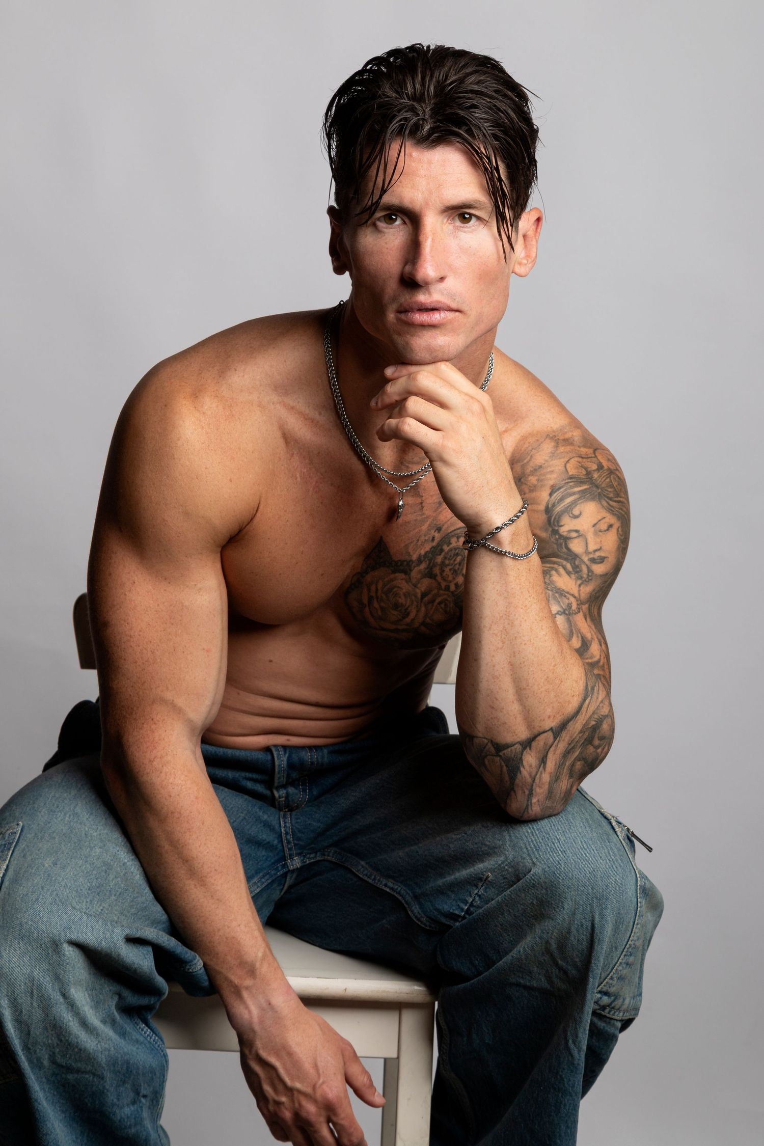 Muscular shirtless man with tattoos sitting on stool in jeans, posing thoughtfully with hand on chin against gray background.