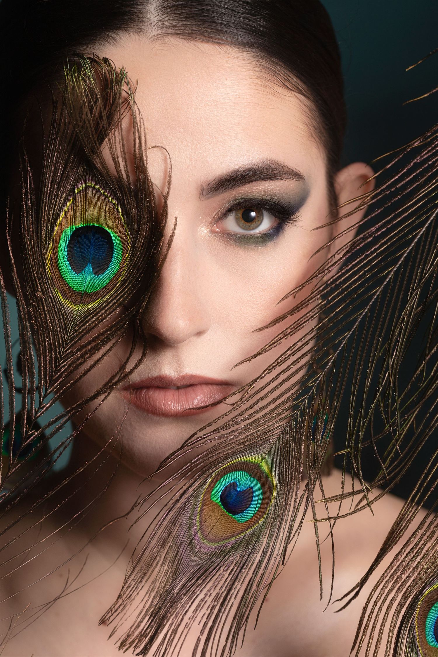 Young woman with dramatic eye makeup poses with vibrant peacock feathers framing her face against dark background.