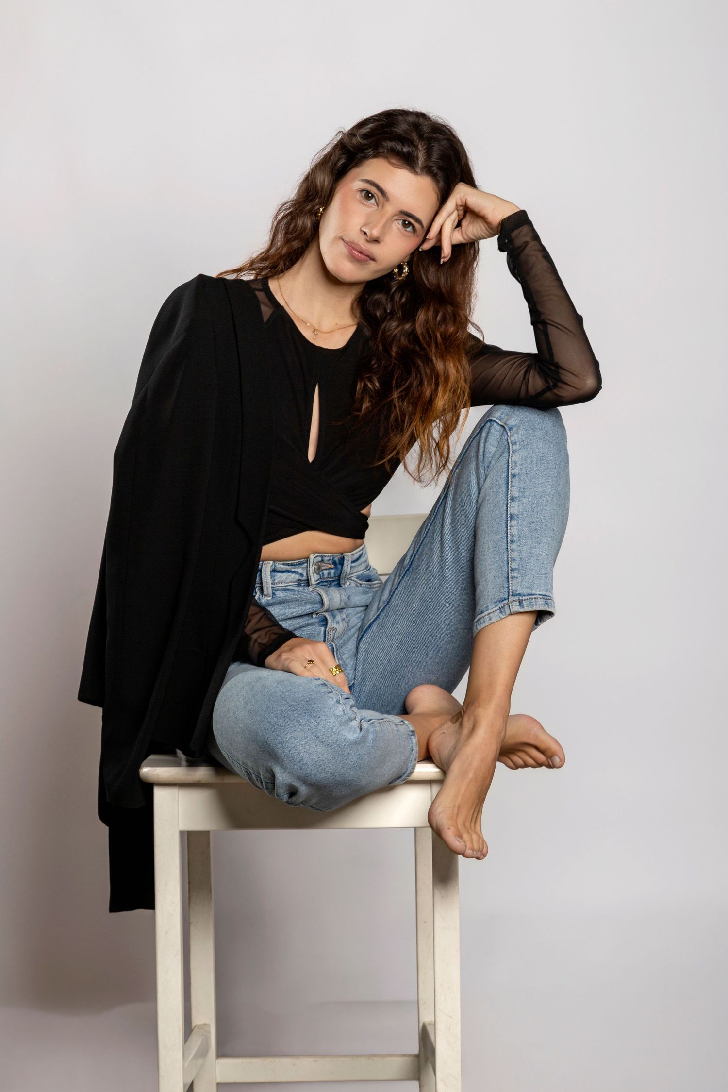 Young woman in black top and ripped jeans sitting casually on a white stool against a light background.