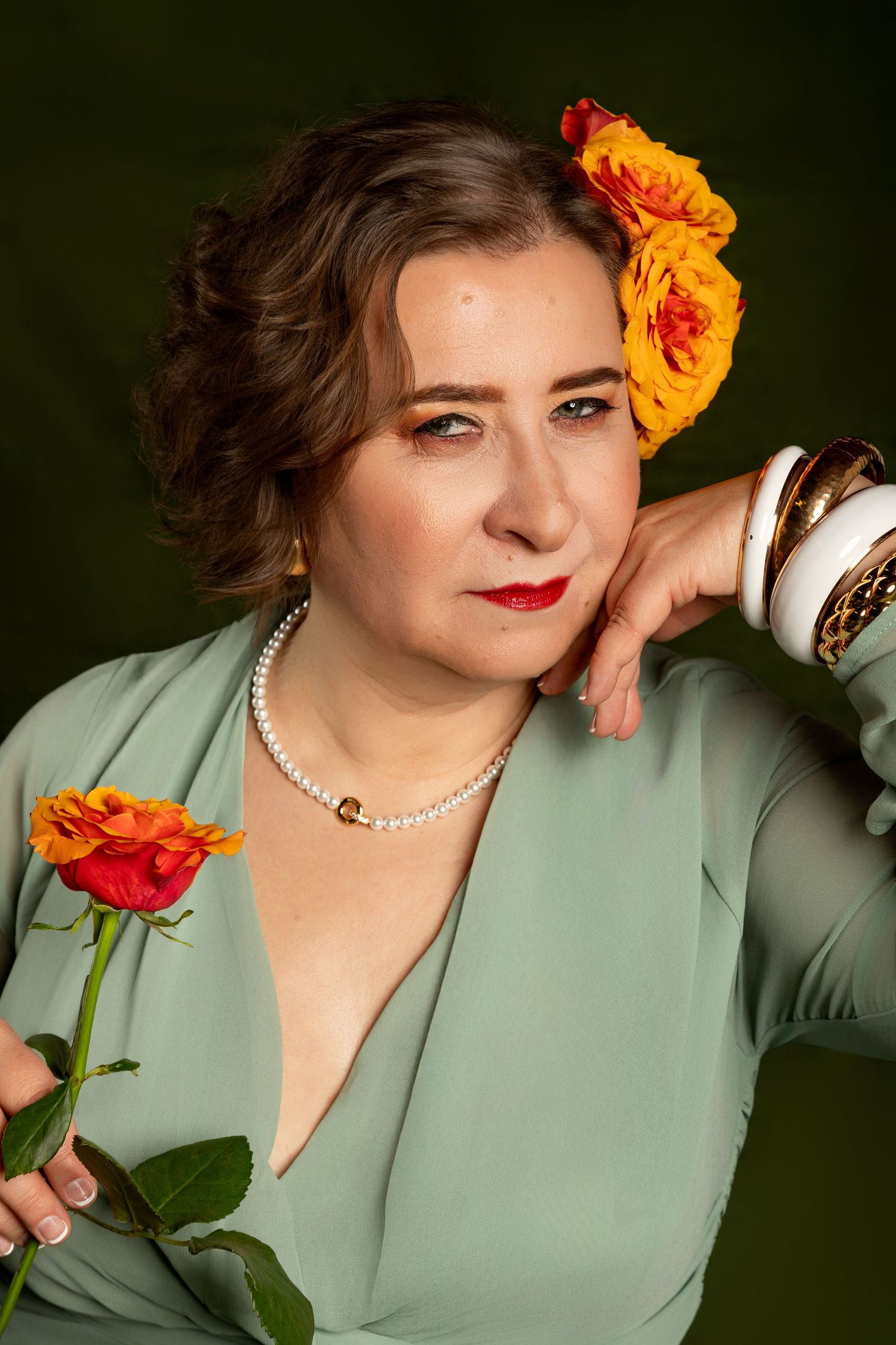 A woman in a mint dress holds an orange rose, wearing pearl necklace and bangles, with roses in her hair.