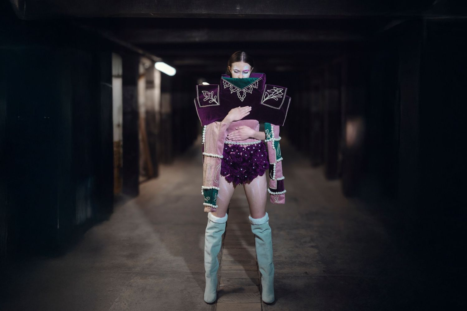 Person in purple avant-garde outfit, pink jacket, white thigh-high boots, and top piece in a dark tunnel.