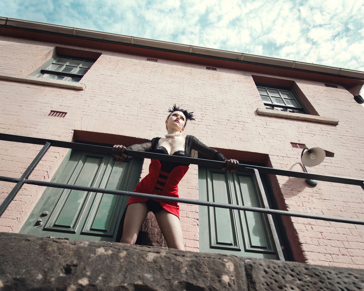 Woman in red corset poses dramatically on a balcony with green shutters against a brick building.