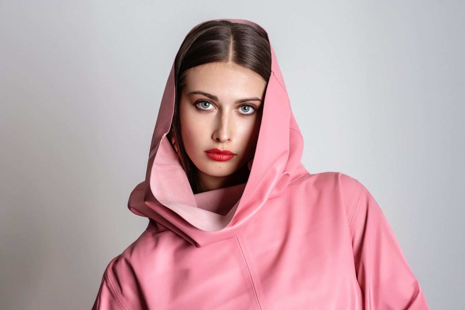 Woman in pink hooded outfit with bold red lips and blue eyes poses against a light gray background.