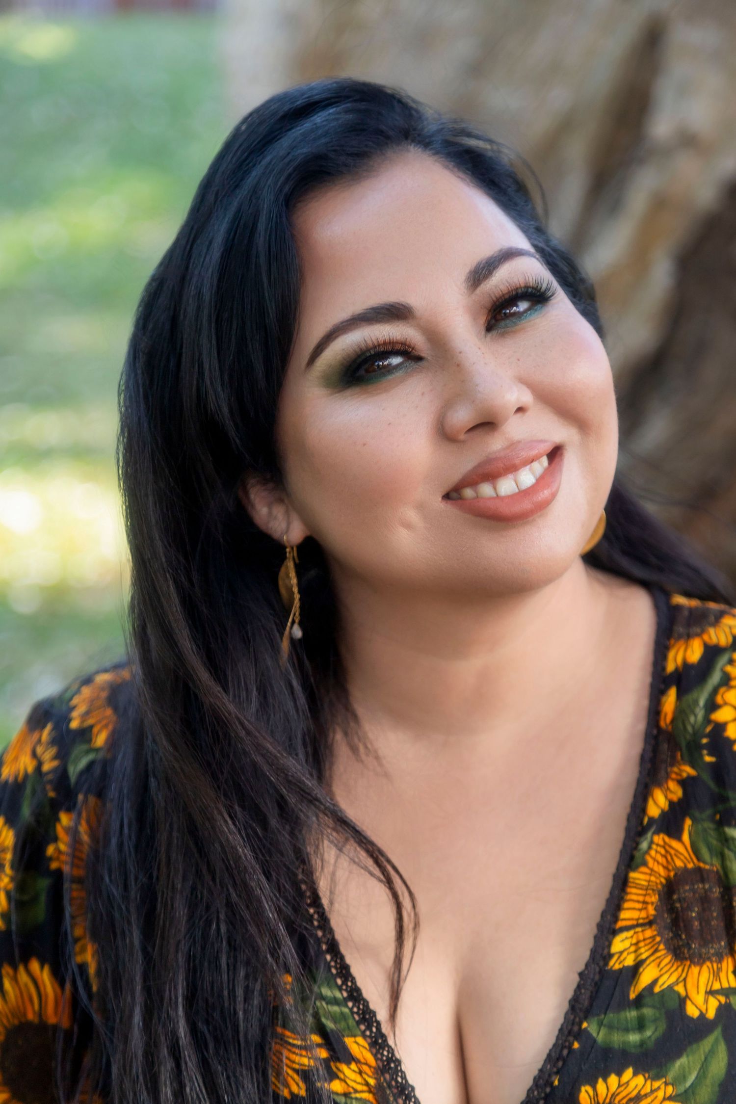 Smiling woman with long black hair wearing a sunflower print top and gold earrings, posing outdoors near a tree.
