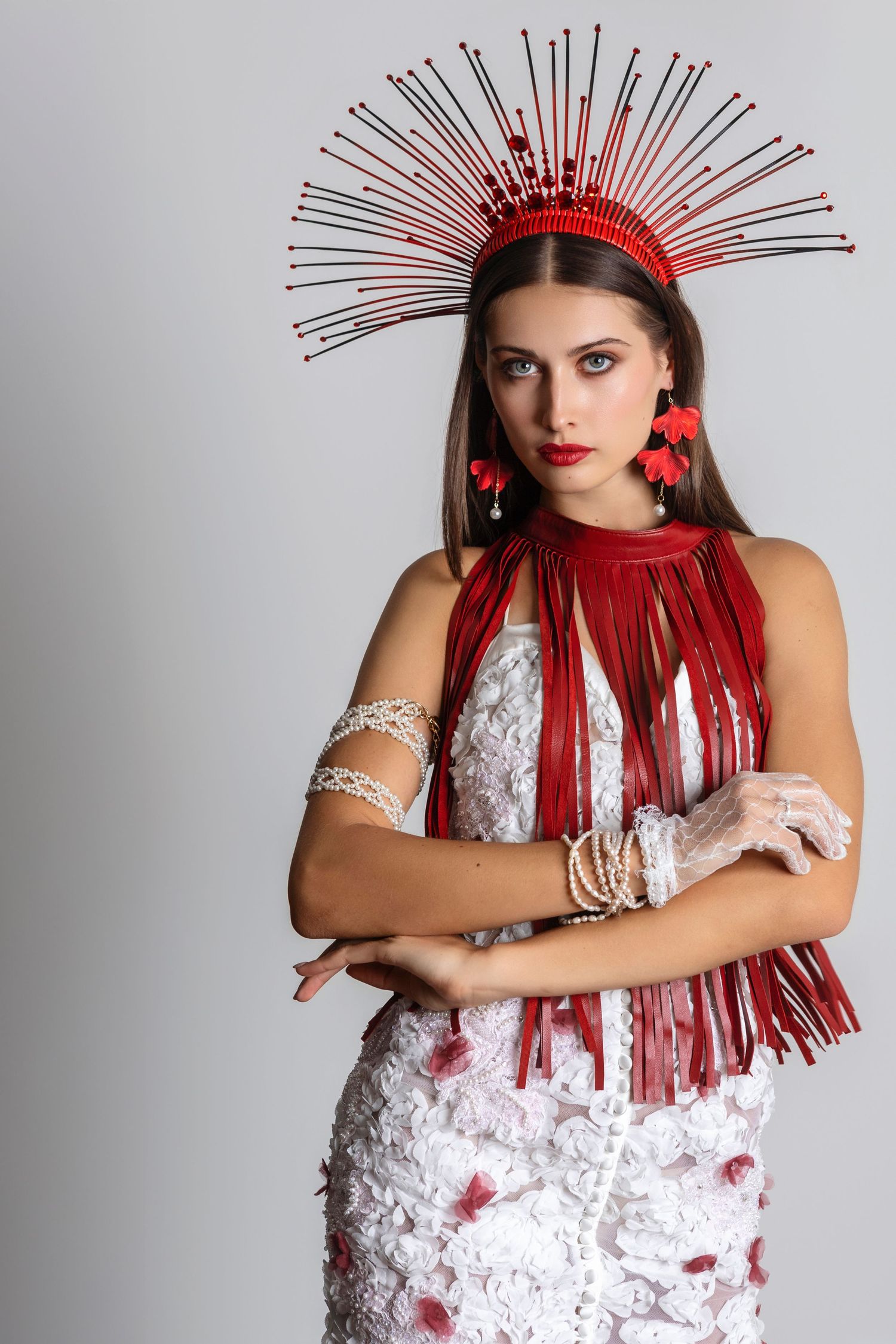 Woman in white dress with red fringe necklace and spiked red crown headpiece, (goddess) poses with arms crossed.